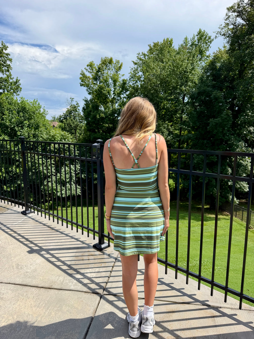 A woman viewed from behind wearing a green and white striped mini dress with thin straps and a crisscross back design, standing on an outdoor balcony with a black metal railing overlooking a green lawn and trees.