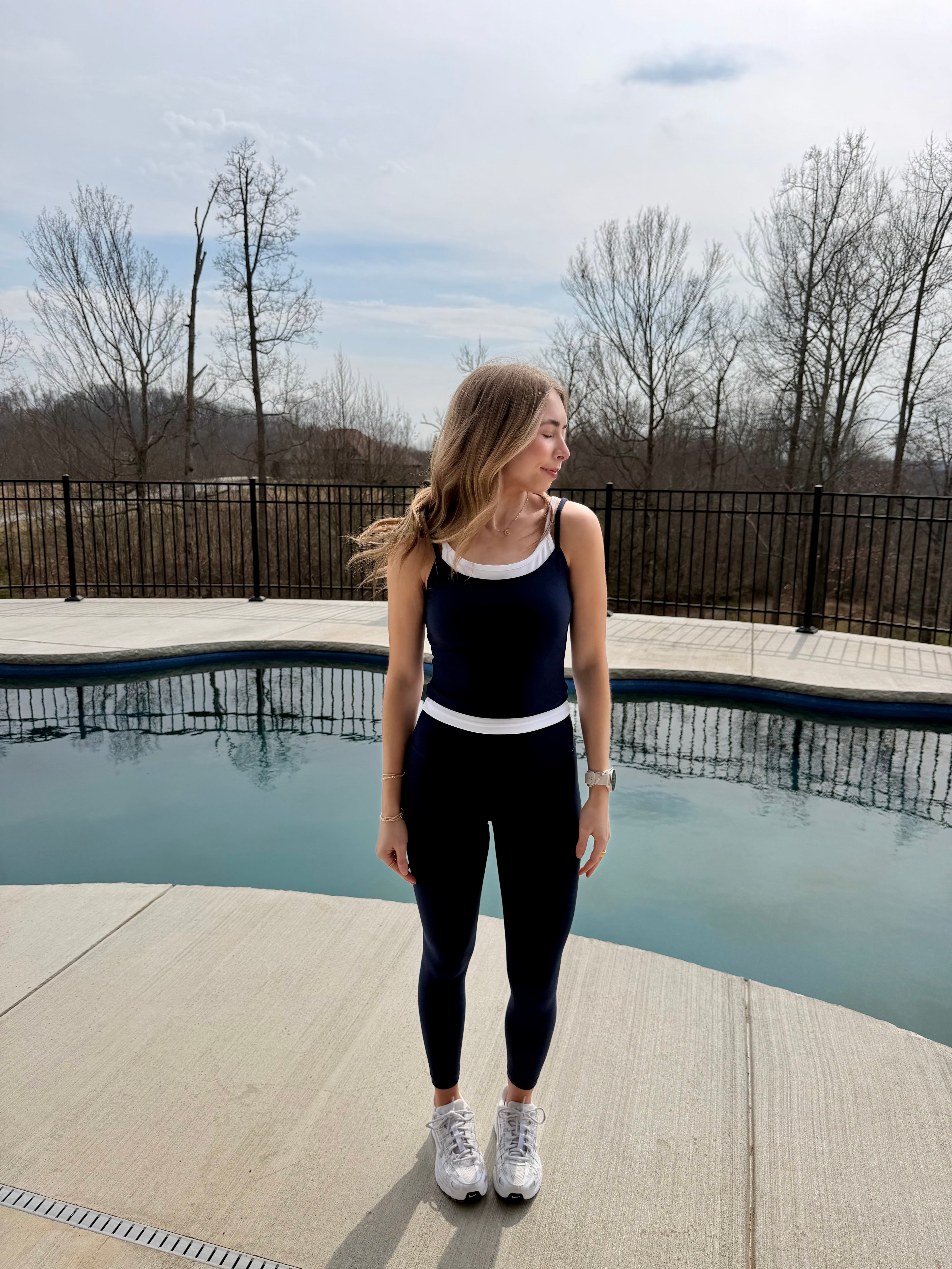 Woman standing by a pool wearing a navy tank top and leggings with white sneakers.