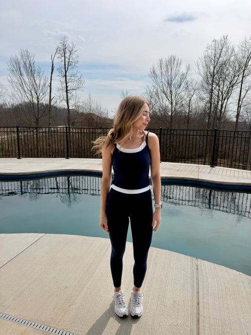 Woman standing by a pool wearing a navy tank top and leggings with white sneakers.