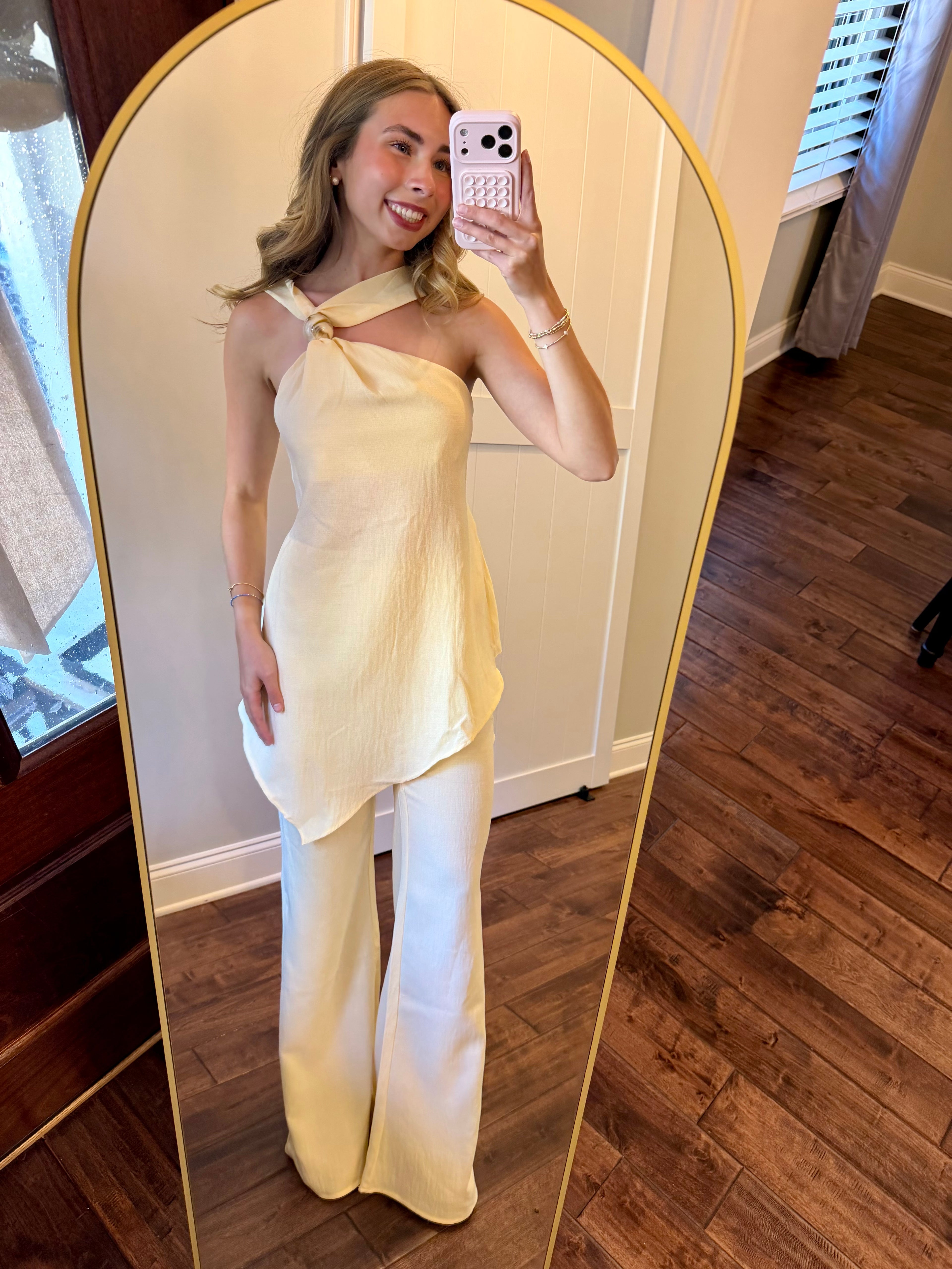 Woman taking a mirror selfie wearing a light-colored outfit in a room with wooden flooring.
