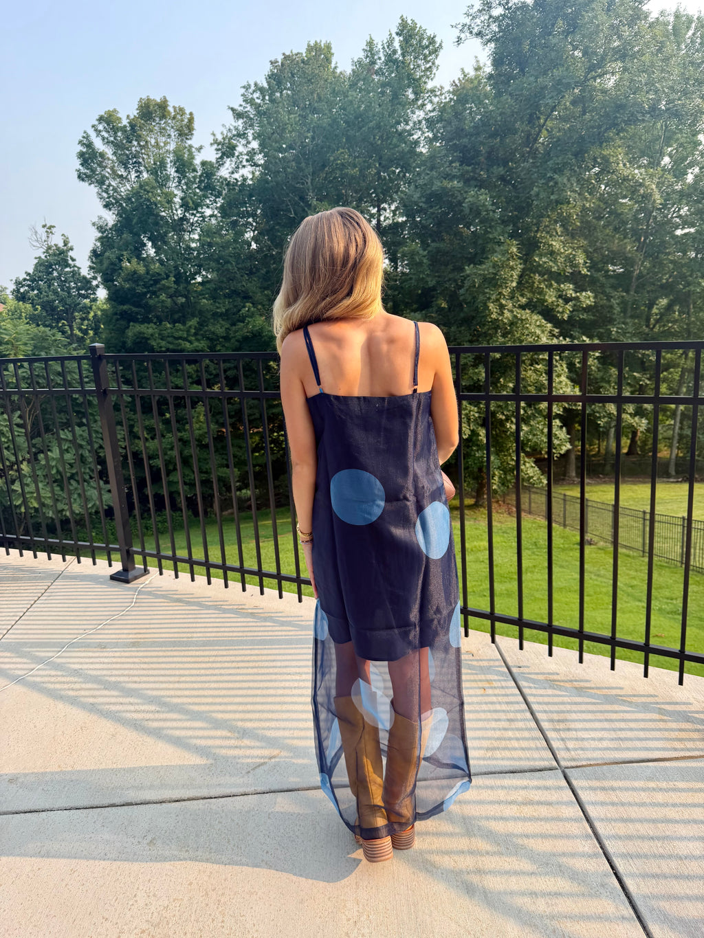 A woman stands on a patio from behind wearing a navy blue sleeveless dress with large light blue and white polka dots, paired with brown knee-high boots, with a black metal railing and green trees in the background.