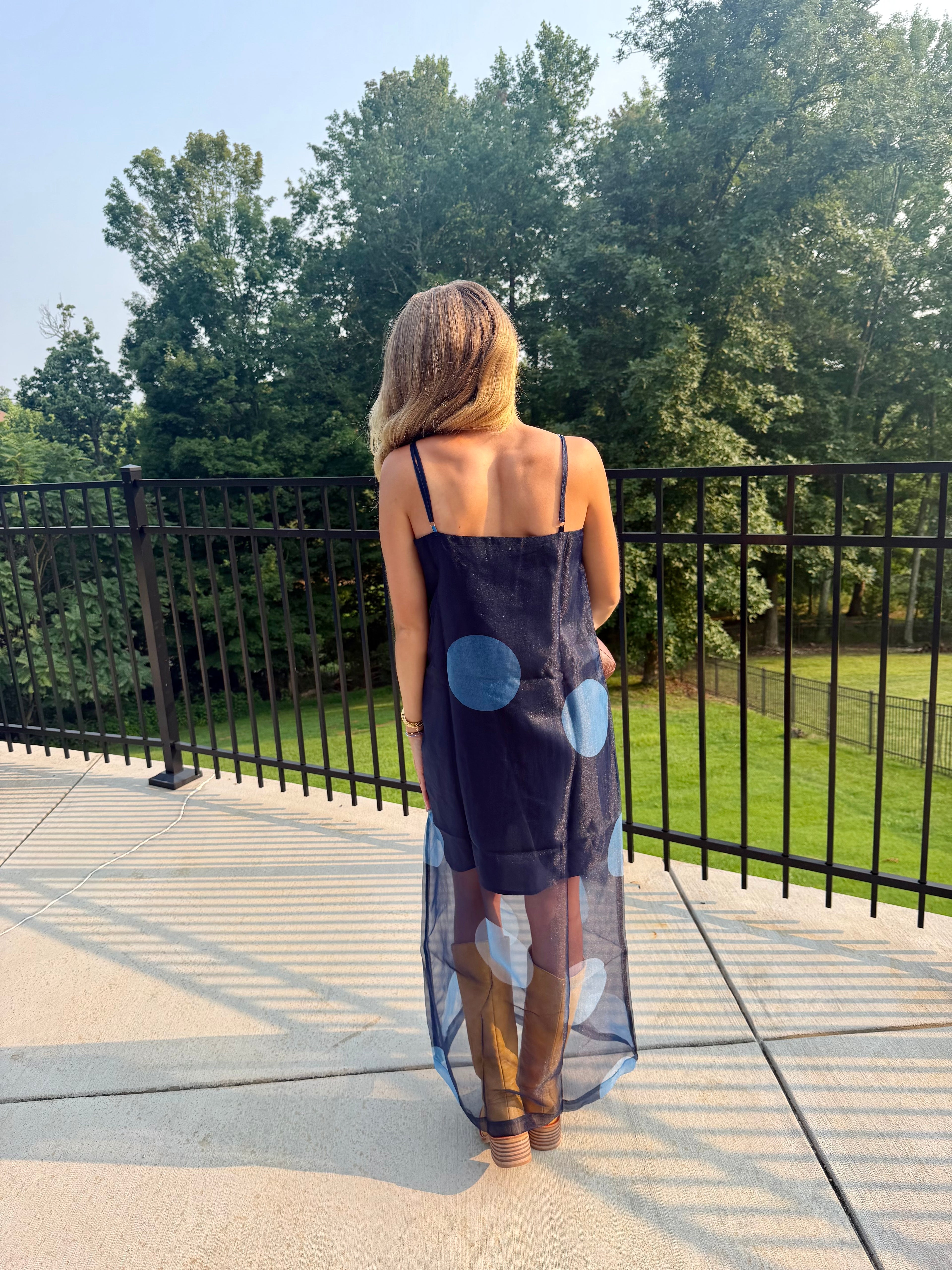 A woman stands on a patio from behind wearing a navy blue sleeveless dress with large light blue and white polka dots, paired with brown knee-high boots, with a black metal railing and green trees in the background.