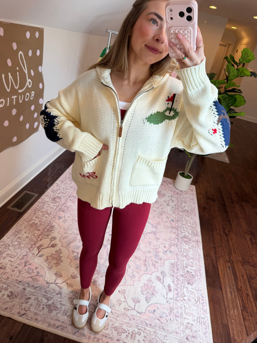 Person wearing a white sweater with green and red patterns, standing on a patterned rug.