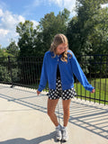 A woman posing outdoors on a concrete walkway wearing a bright blue varsity-style jacket, black and white polka dot shorts, a black top, and white sneakers, with trees and a fence in the background.