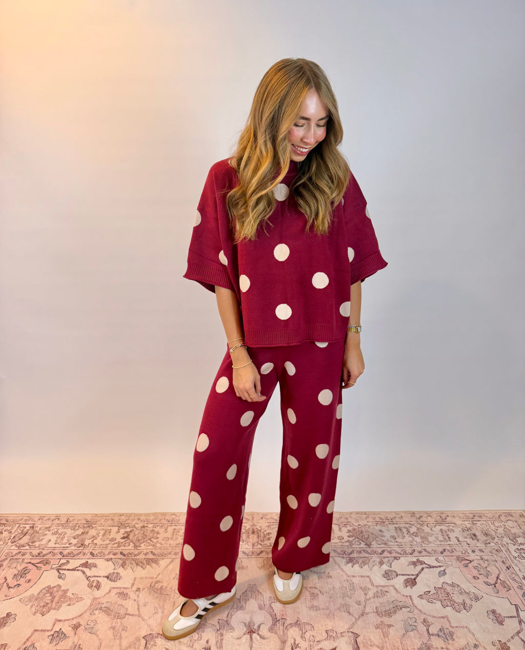 A woman modeling a matching two-piece lounge set in deep burgundy with large white polka dots, consisting of a short-sleeve cropped top and wide-leg pants, paired with white sneakers.