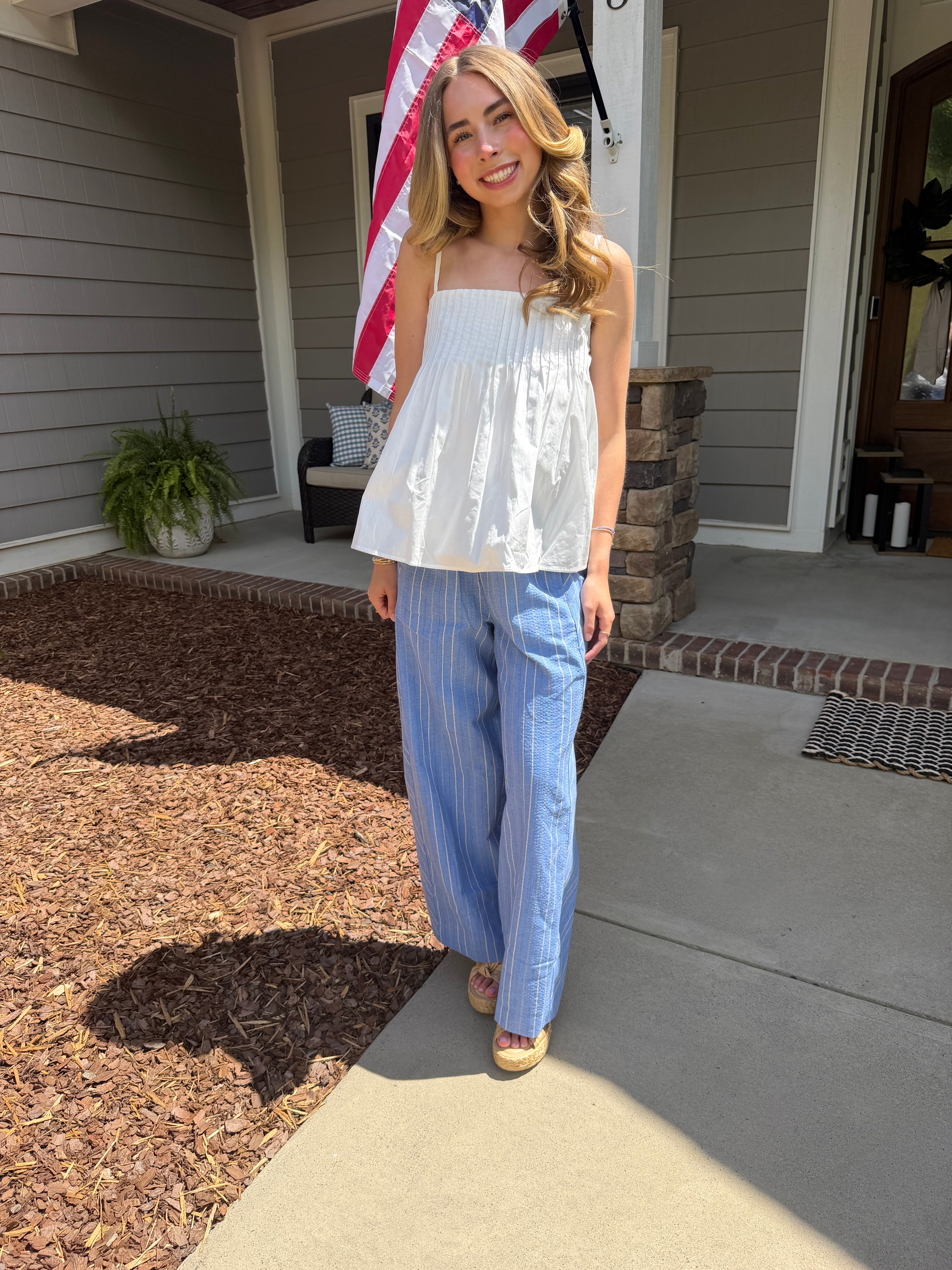 Woman standing on a porch wearing a white top and blue striped pants, with an American flag in the background.