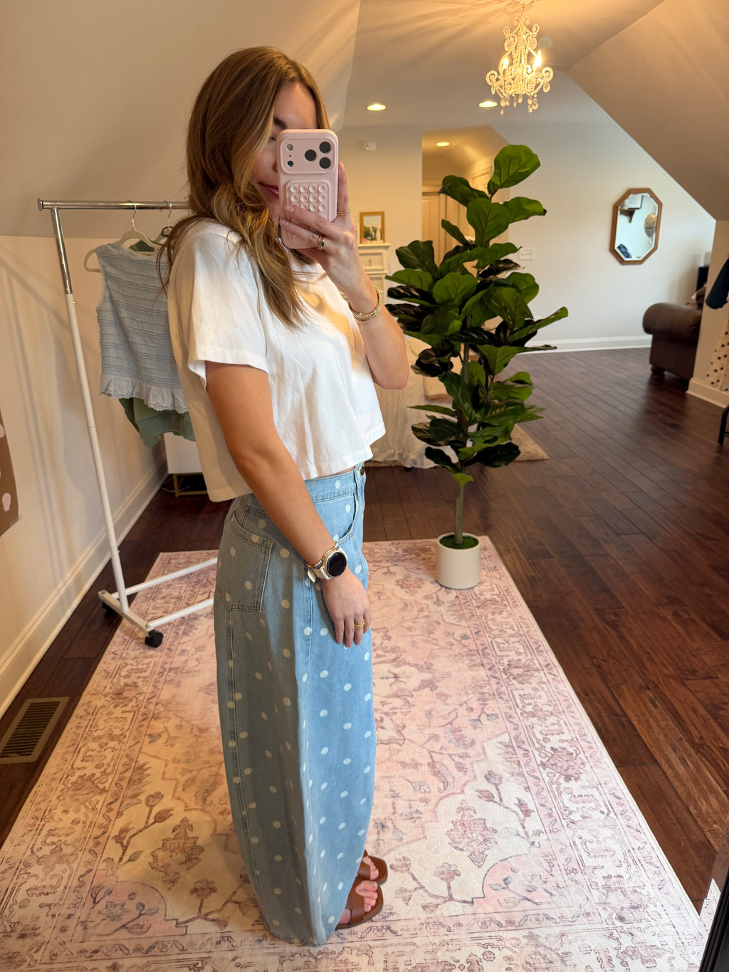 Woman taking a mirror selfie wearing a white top and light blue pants with a polka dot pattern in a room with a plant and rug.