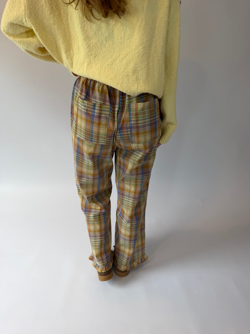 A person shown from the back wearing loose yellow knit top and plaid pants in beige, blue, and brown tones, standing against a plain light gray studio background.
