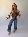 A young woman modeling a pink and brown striped knit sweater, paired with wide-leg blue jeans and pink flat shoes, standing against a light gray studio background.