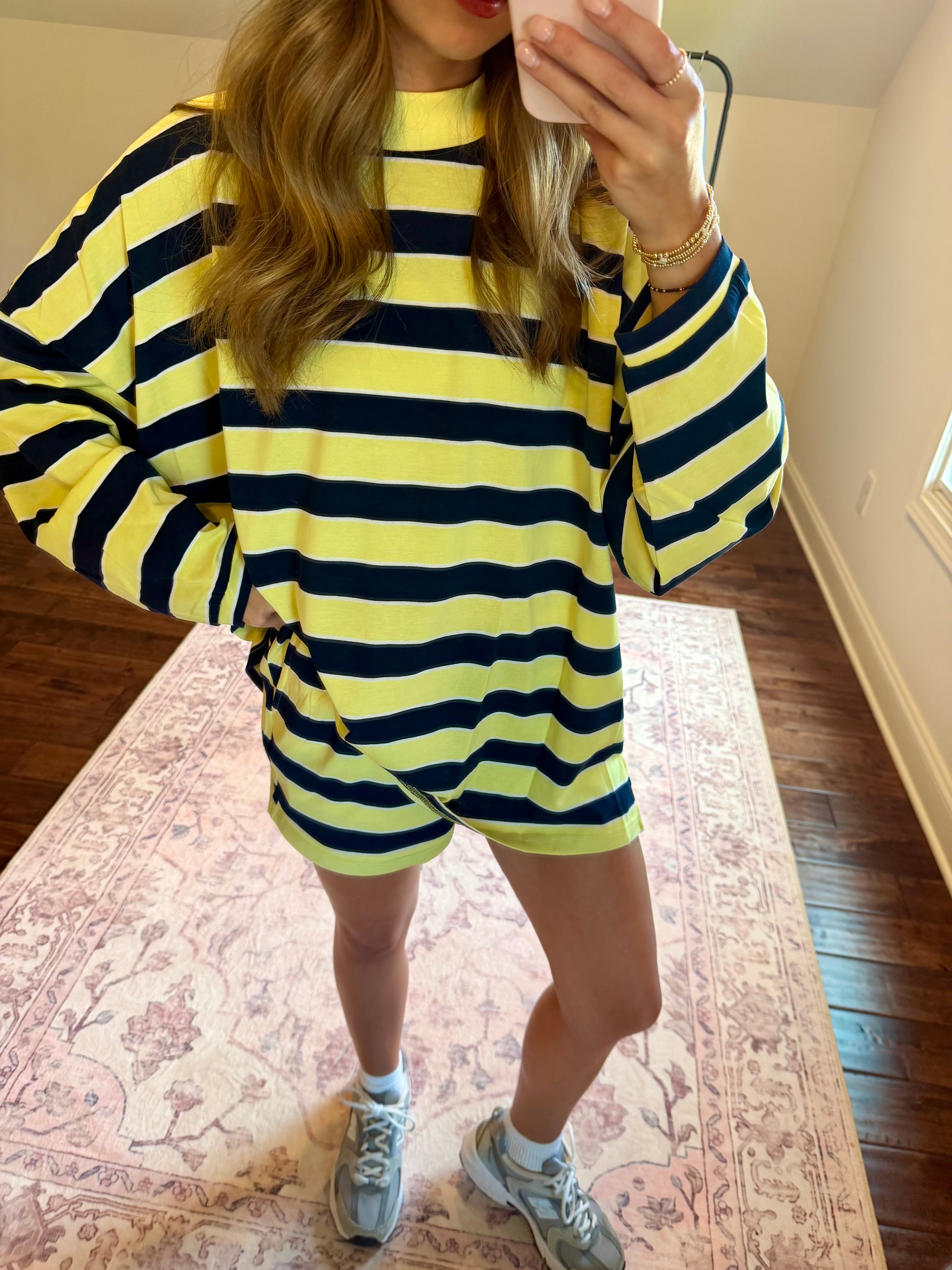 A woman takes a mirror selfie wearing a yellow and navy blue horizontally striped long-sleeve knit top and matching shorts set, paired with gray sneakers, standing on a patterned rug in a room with hardwood floors.