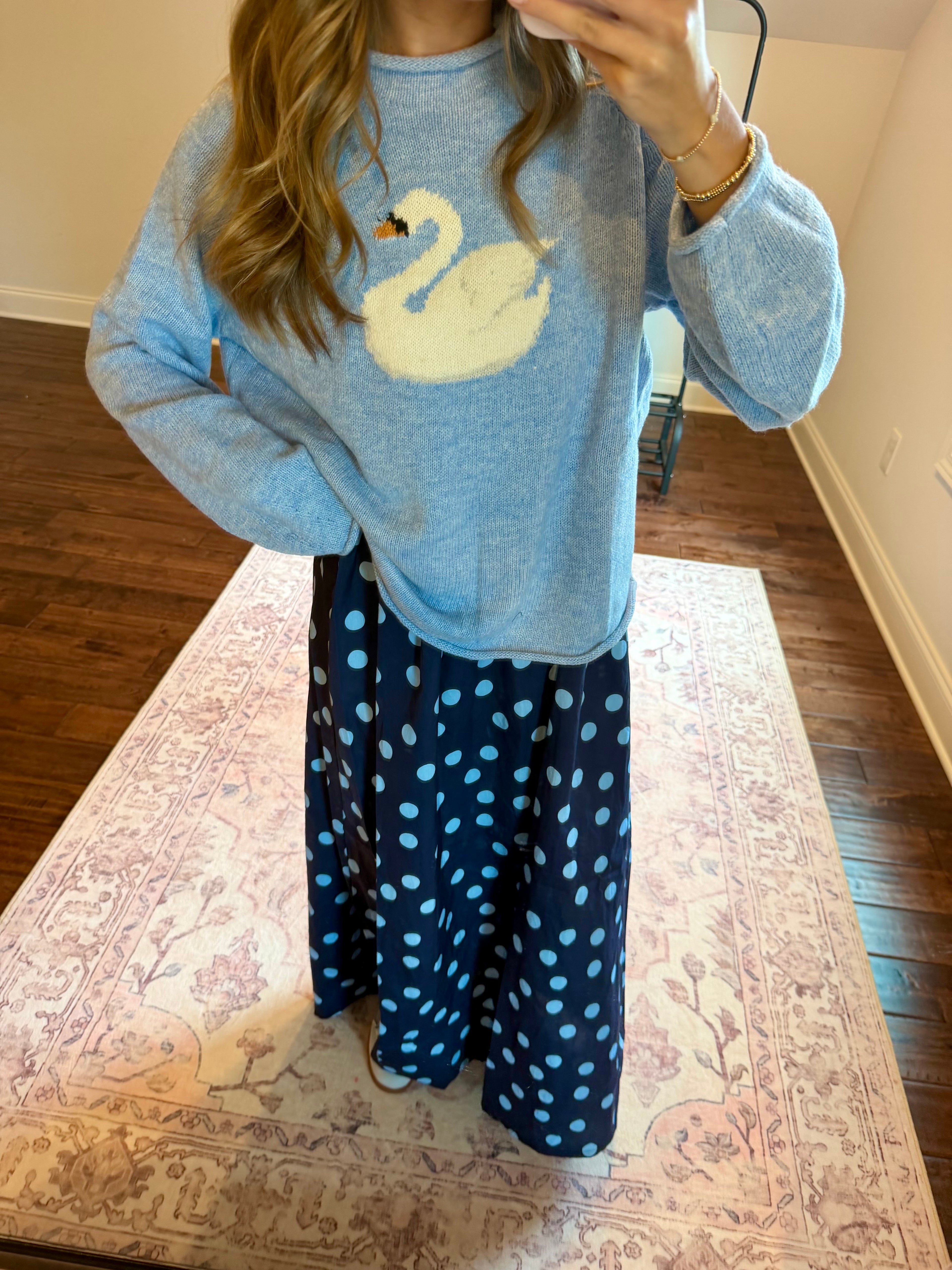 A person wearing a light blue long-sleeve sweater with a white swan graphic on the front, paired with a navy blue maxi skirt with light blue polka dots, standing in a room with hardwood floors and a patterned rug.