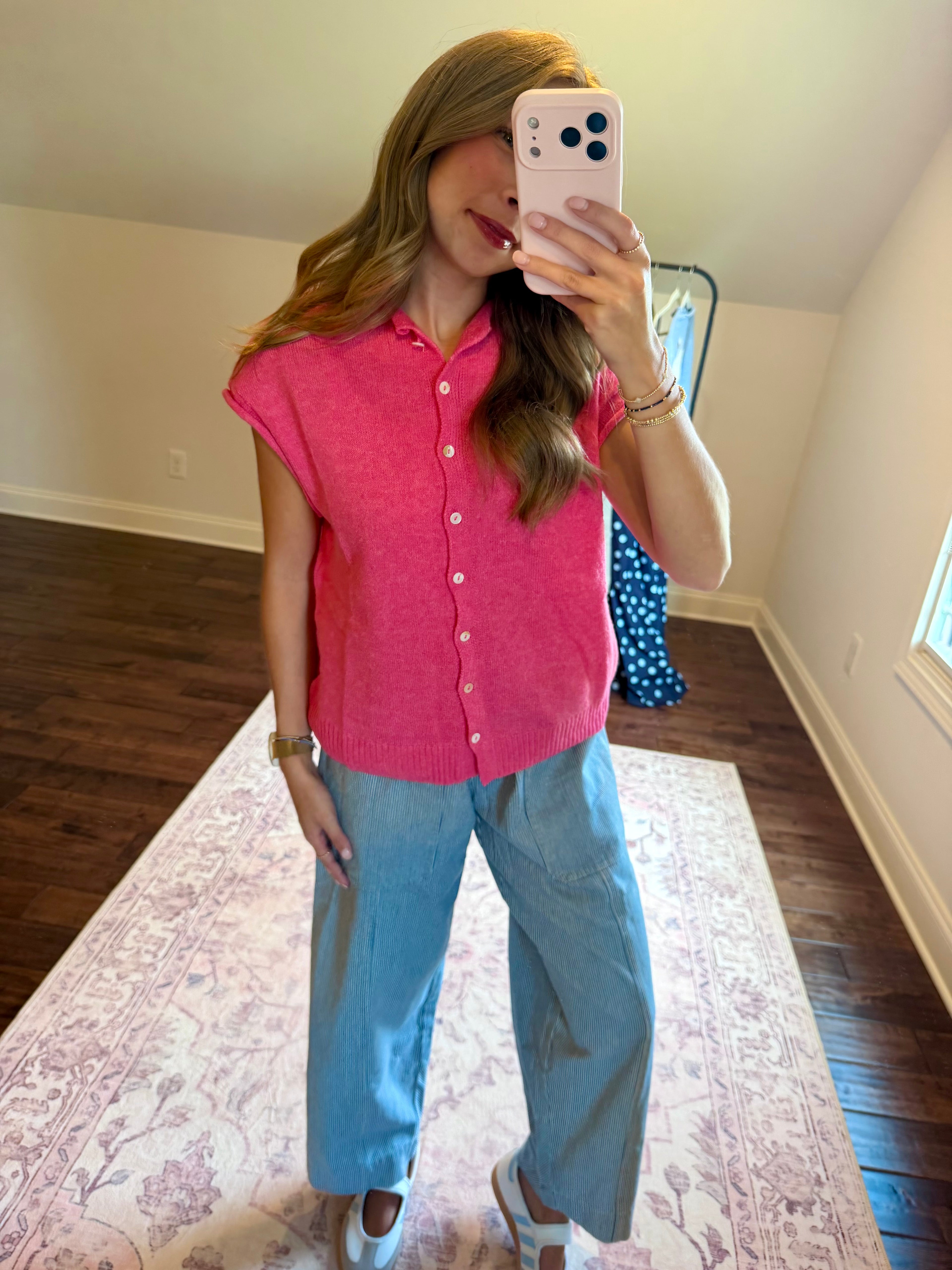 A woman takes a mirror selfie in a room, wearing a bright pink short-sleeve button-up shirt and light blue wide-leg jeans.