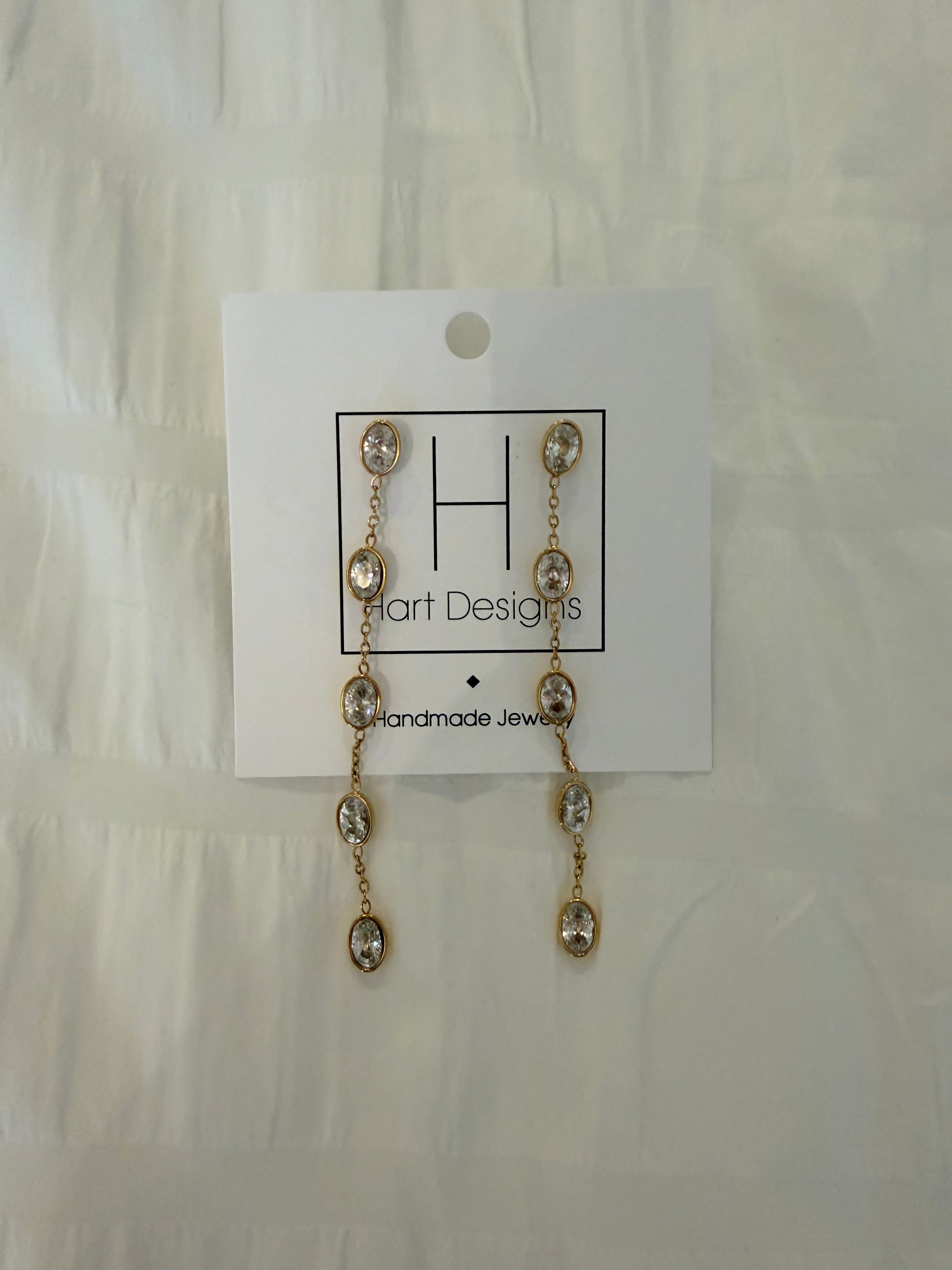 Gold earrings with clear stones on a white card with 'Hart Designs' branding, placed on a textured beige surface.