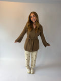 A woman modeling a brown long-sleeve belted wrap top with cream pants featuring brown polka dots, standing against a plain light gray studio backdrop.