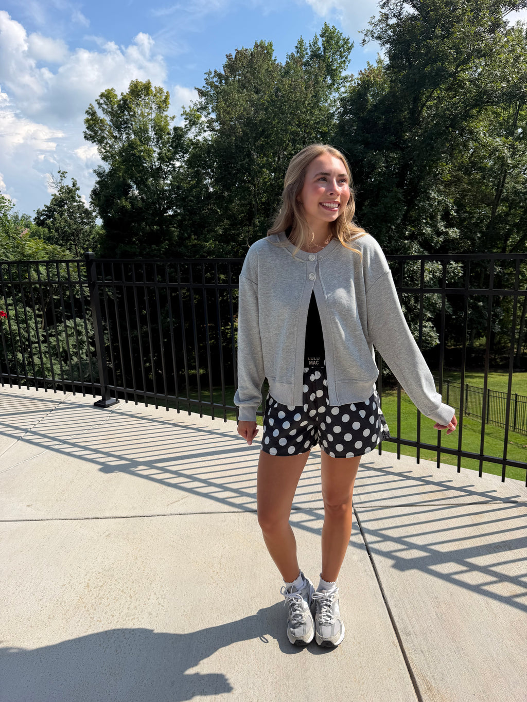 A woman stands outdoors on a concrete walkway wearing a light gray zip-up hoodie, black and white polka dot shorts, a black top underneath, and white sneakers. Trees and a black metal fence are visible in the background under a blue sky.