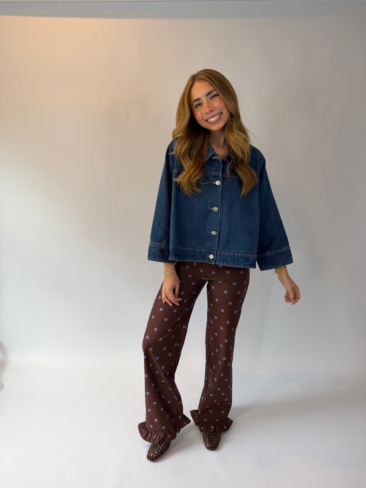 A woman modeling a dark blue cropped denim jacket and brown wide-leg pants with a small white polka dot pattern, standing against a plain light gray studio background.