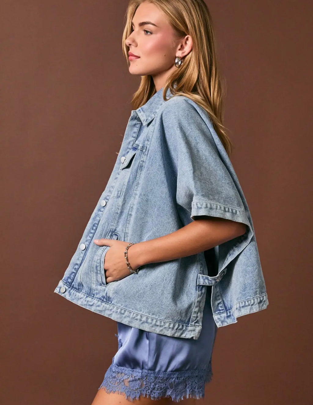 A woman wearing a light blue oversized denim jacket with short sleeves, side pockets, and a button front, paired with matching denim shorts, posed against a solid brown studio background.