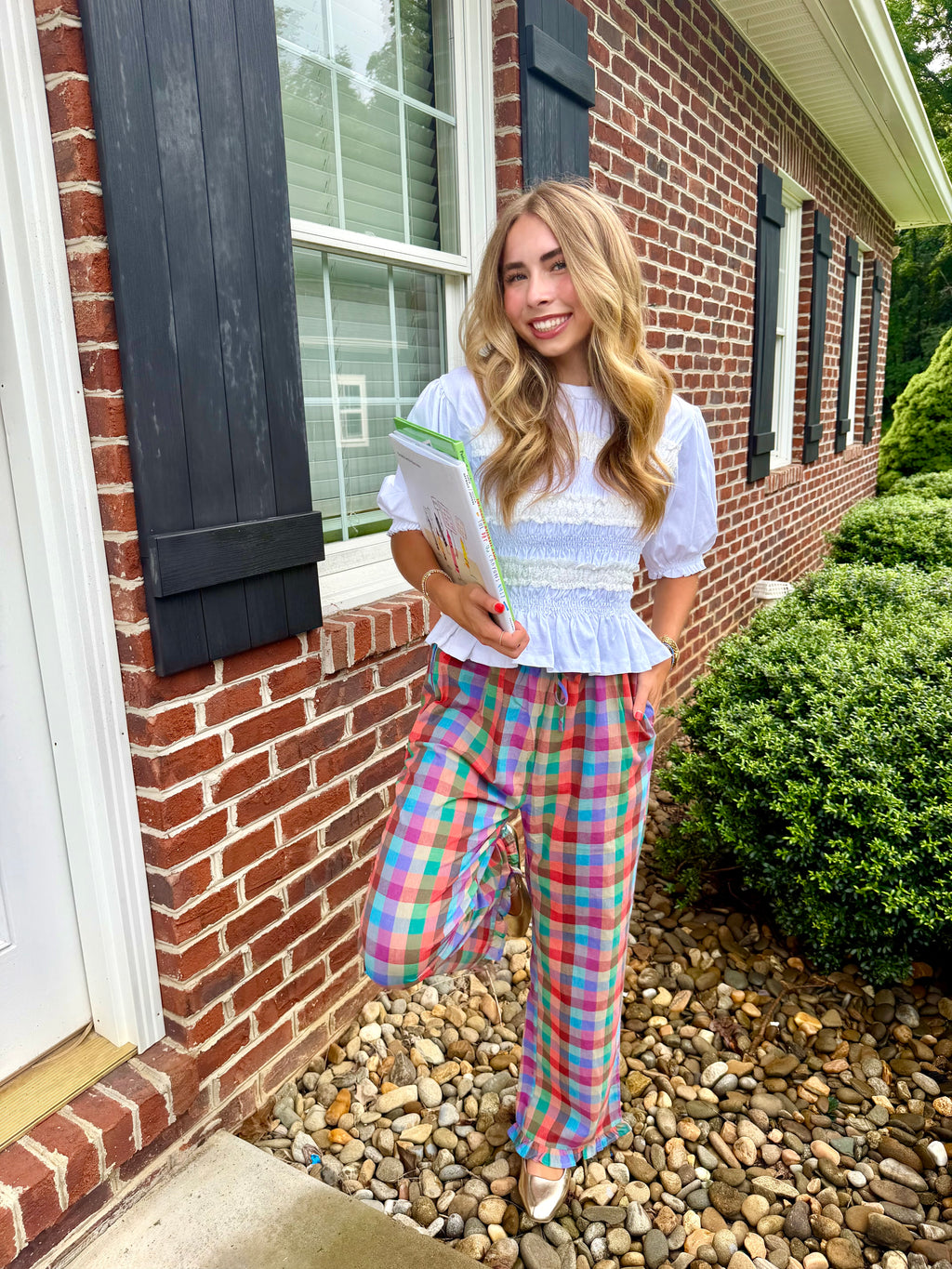 A woman stands outdoors in front of a brick house wearing a white ruffled short-sleeve top and bright multicolor plaid pants, holding a book or magazine.