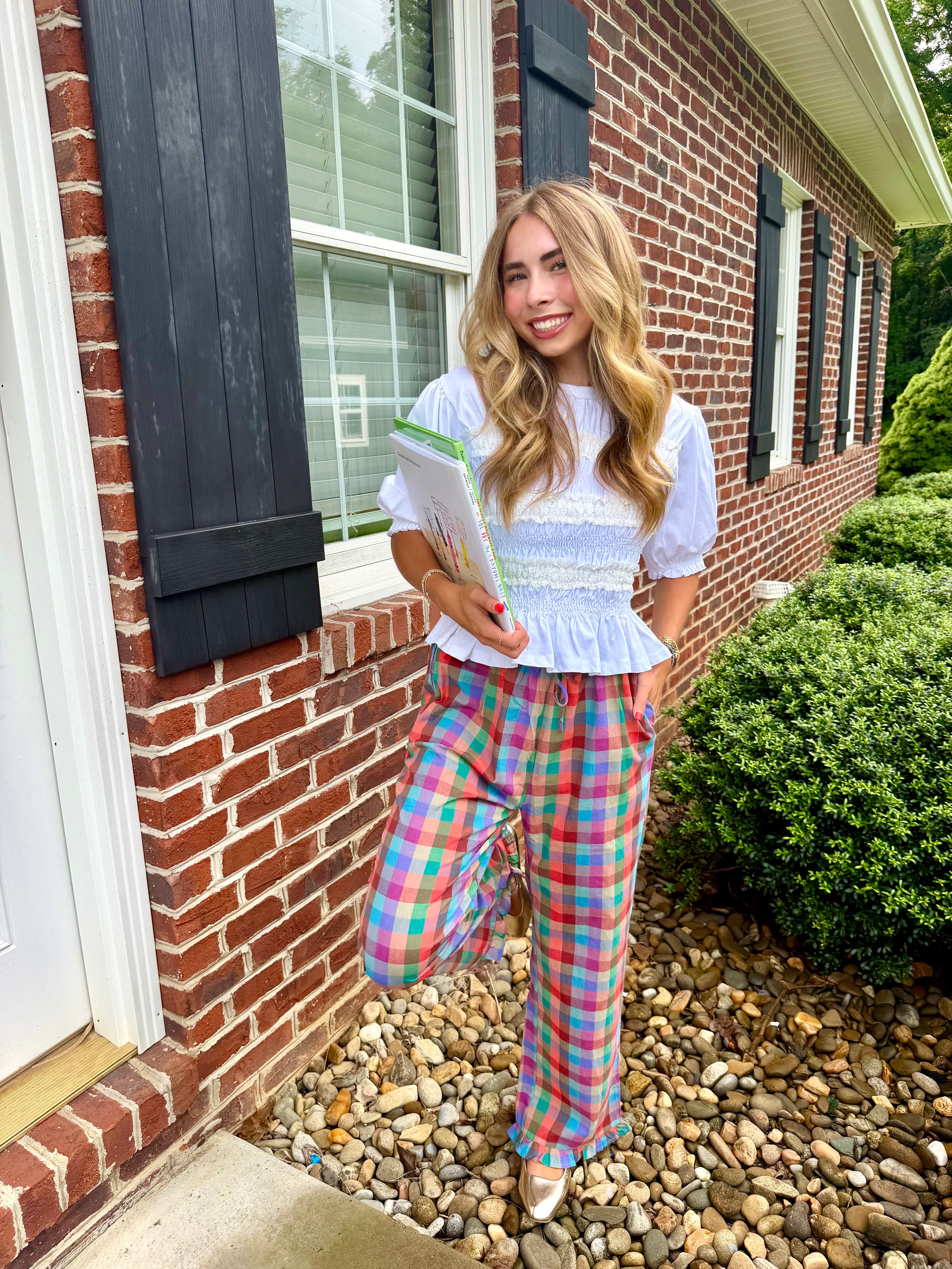 A woman stands outdoors in front of a brick house wearing a white ruffled short-sleeve top and bright multicolor plaid pants, holding a book or magazine.