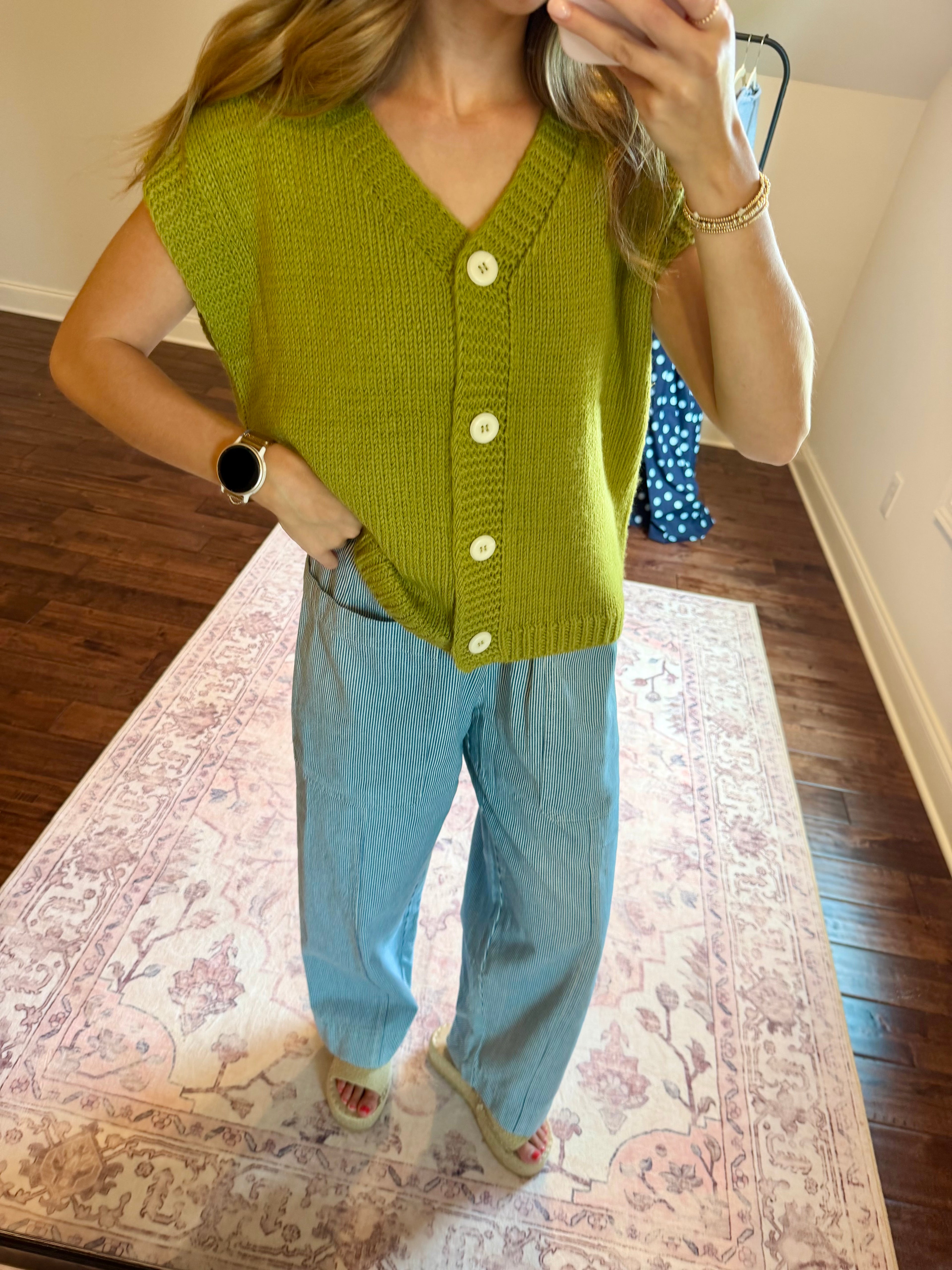 A woman takes a mirror selfie wearing a sleeveless olive green knit cardigan with white buttons and light blue wide-leg jeans, standing on a patterned rug in a room with hardwood floors.