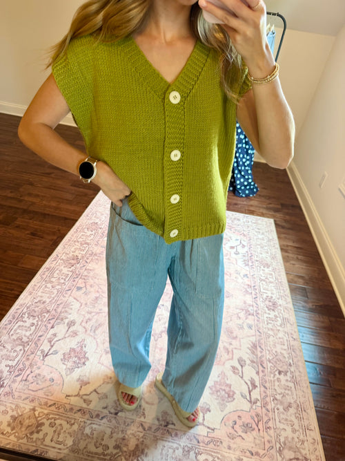 A woman takes a mirror selfie wearing a sleeveless olive green knit cardigan with white buttons and light blue wide-leg jeans, standing on a patterned rug in a room with hardwood floors.