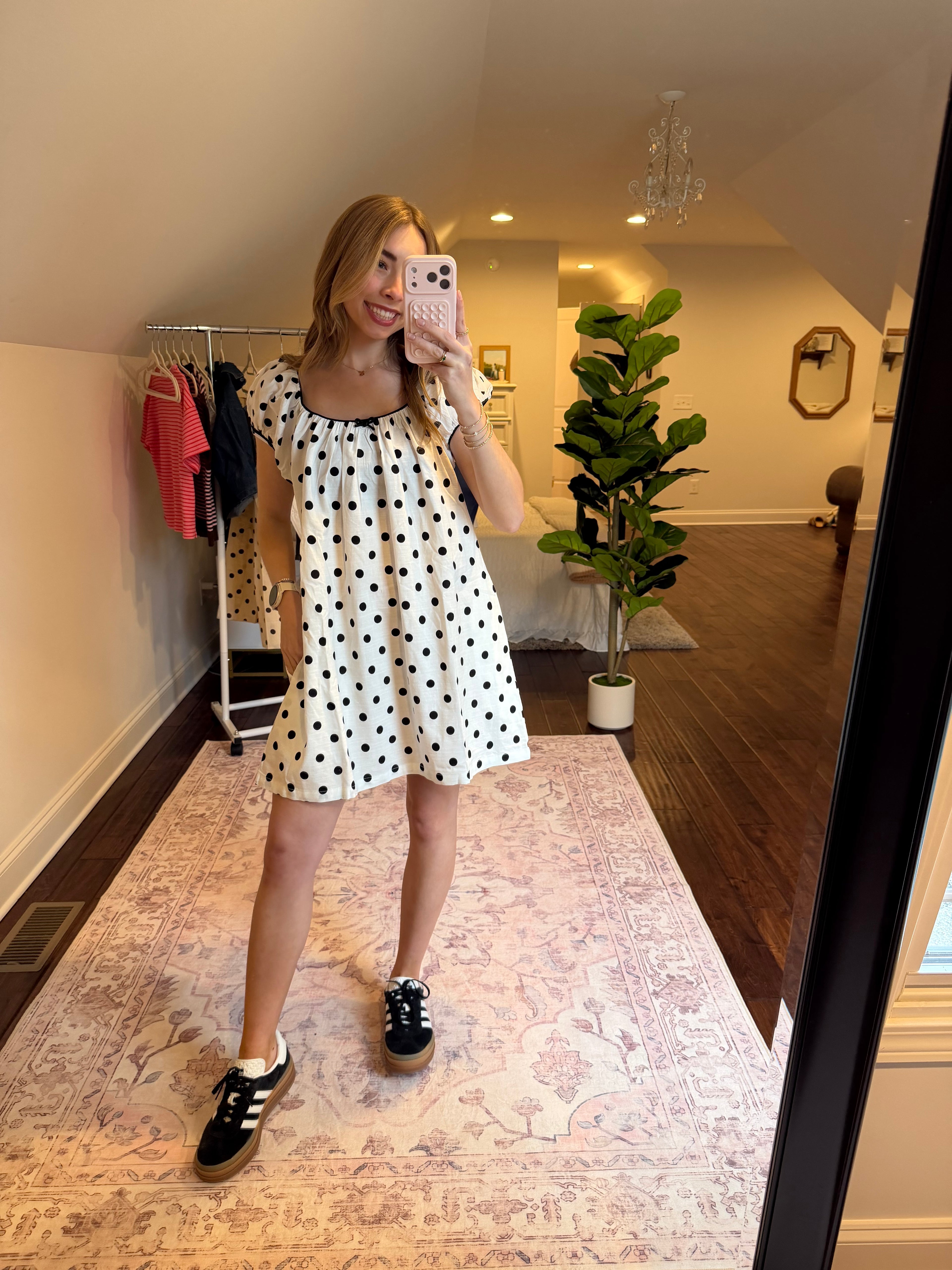Woman in a polka dot dress standing in a room with a plant and patterned rug.
