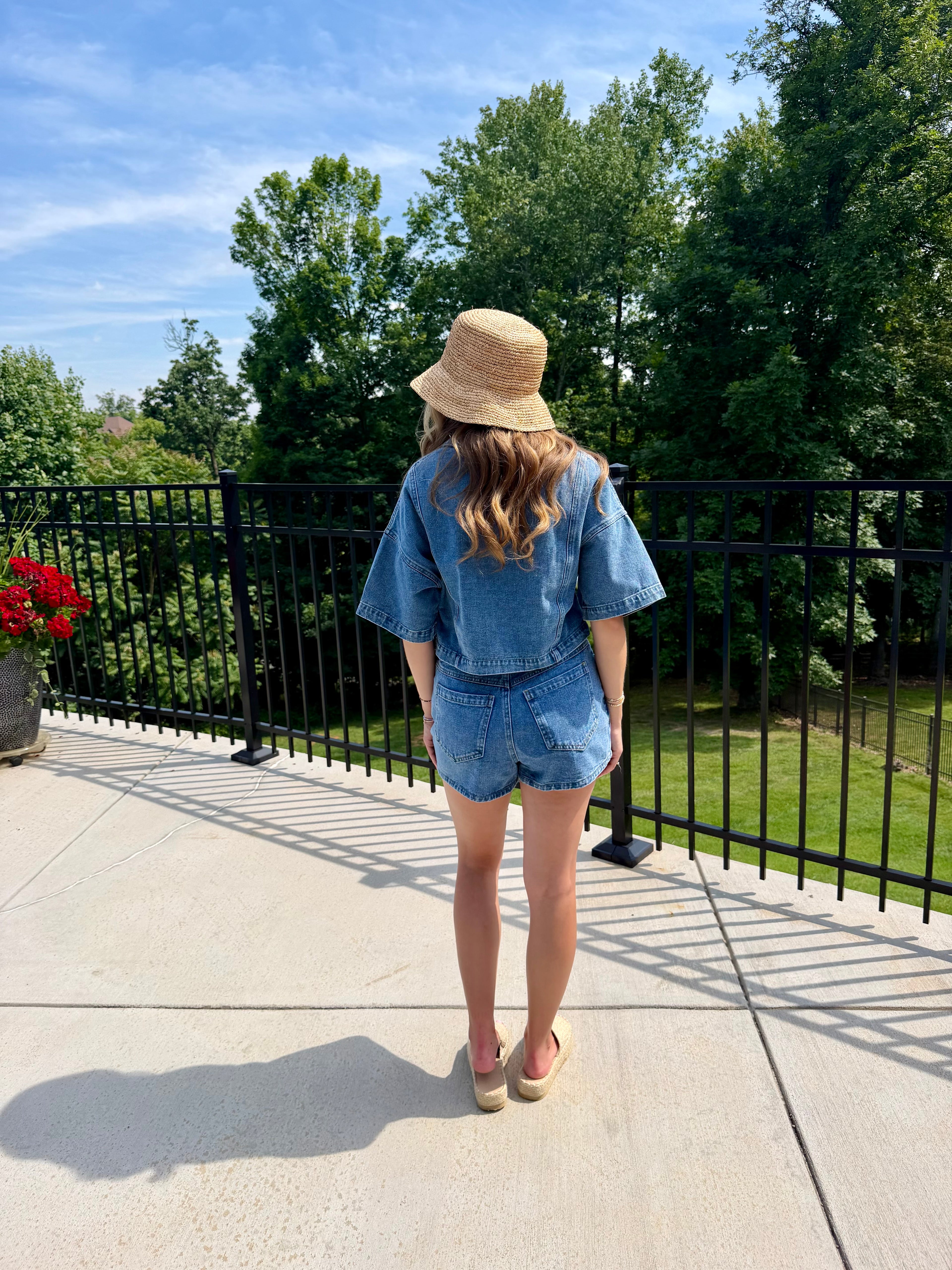 Person wearing a denim outfit and straw hat standing on a patio with trees in the background