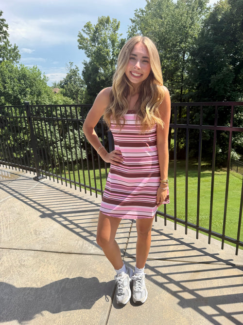 A young woman stands outdoors on a concrete walkway wearing a pink and brown striped tube dress with white sneakers. Trees and a black metal fence are visible in the background.