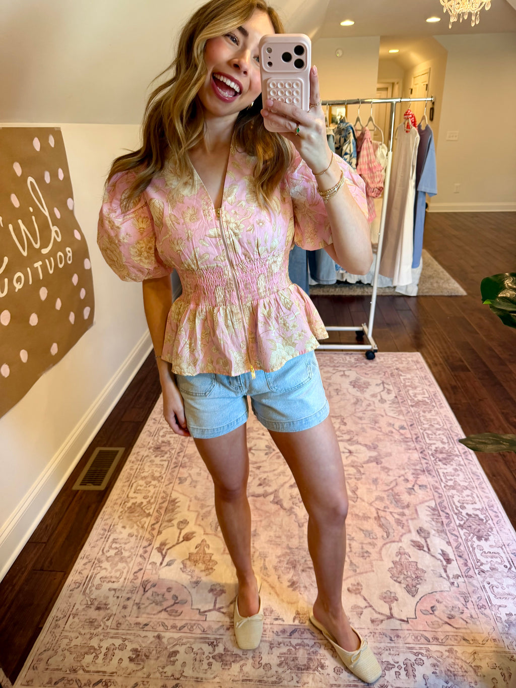 Woman taking a mirror selfie wearing a floral top and denim shorts in a home setting.