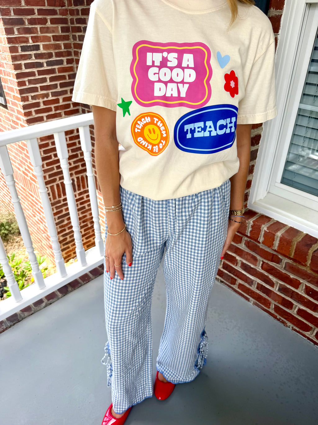 A person standing on a porch wearing a cream graphic T-shirt with colorful school-themed text and patches, blue-and-white gingham wide-leg pants, and red shoes.