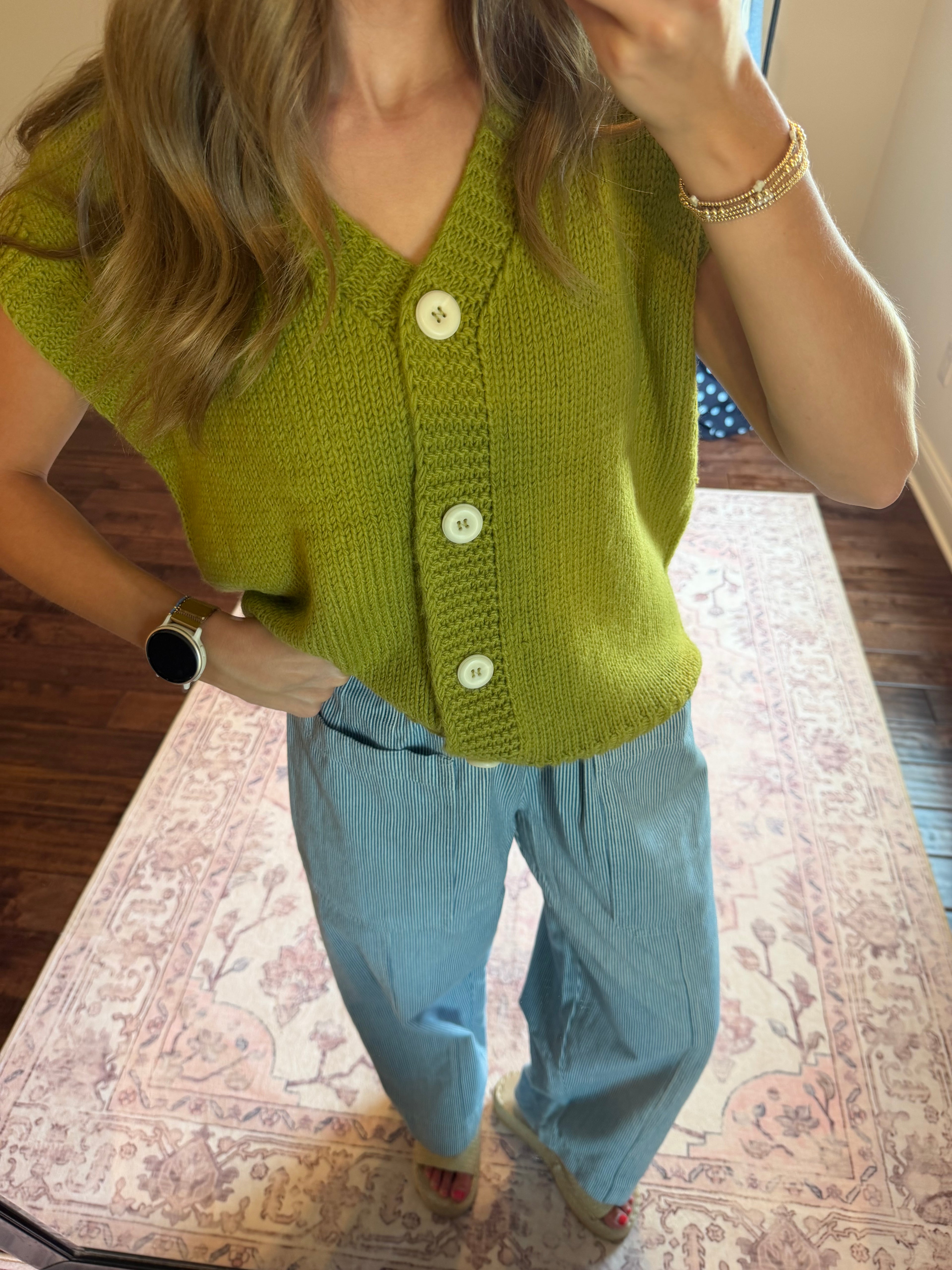 A woman wearing a lime green knit cardigan vest with white buttons and light blue wide-leg pants, standing on a patterned rug in a home setting.