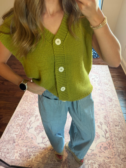 A woman wearing a lime green knit cardigan vest with white buttons and light blue wide-leg pants, standing on a patterned rug in a home setting.