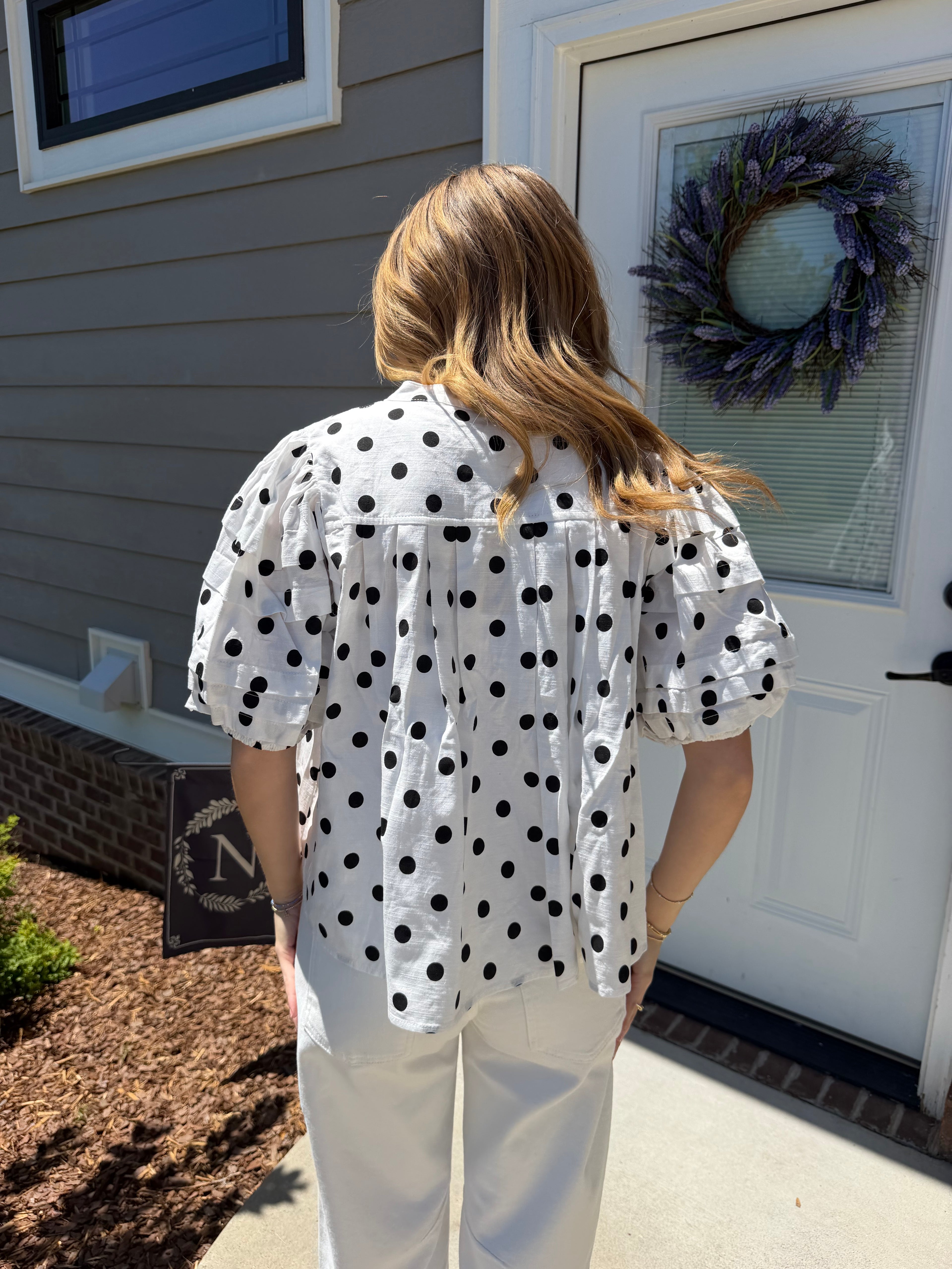 Person wearing a polka dot blouse standing in front of a house with a wreath on the door.