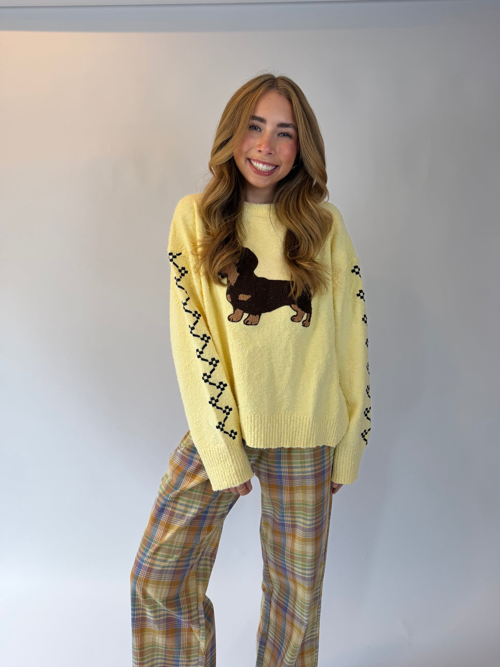 A woman modeling a pale yellow knit sweater with a brown dachshund graphic on the chest and black zigzag knit patterns along the sleeves, paired with plaid pants against a plain light gray studio background.