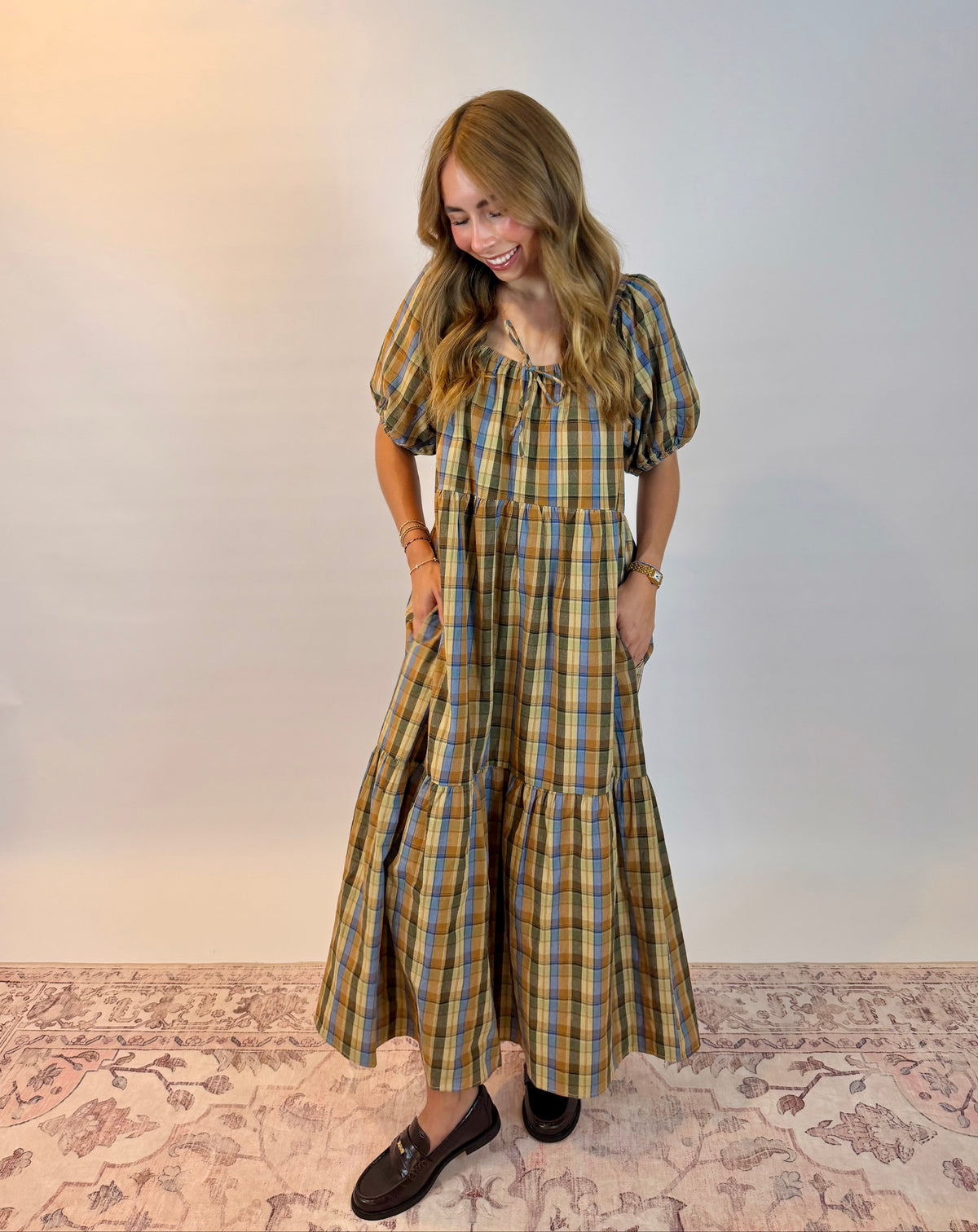 A woman modeling a yellow, green, and blue plaid maxi dress with puff sleeves, standing on a patterned rug against a plain light gray wall.