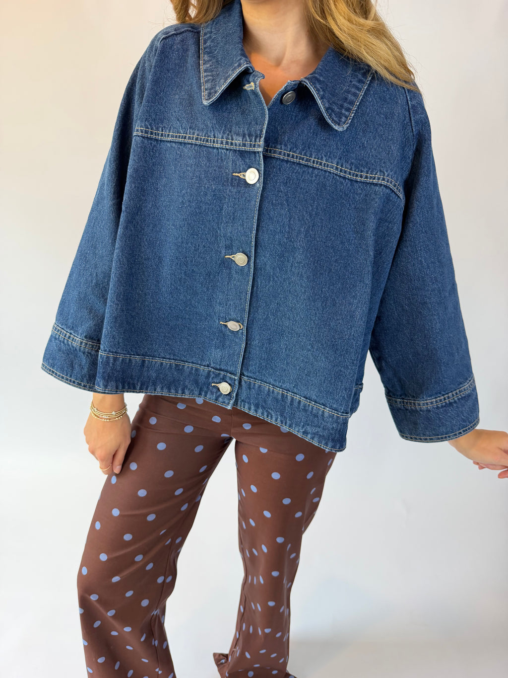 A woman wearing a cropped blue denim jacket with silver buttons and brown pants with light blue polka dots, photographed against a plain light background.