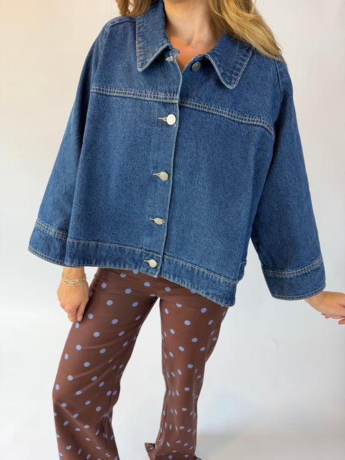 A woman wearing a cropped blue denim jacket with silver buttons and brown pants with light blue polka dots, photographed against a plain light background.