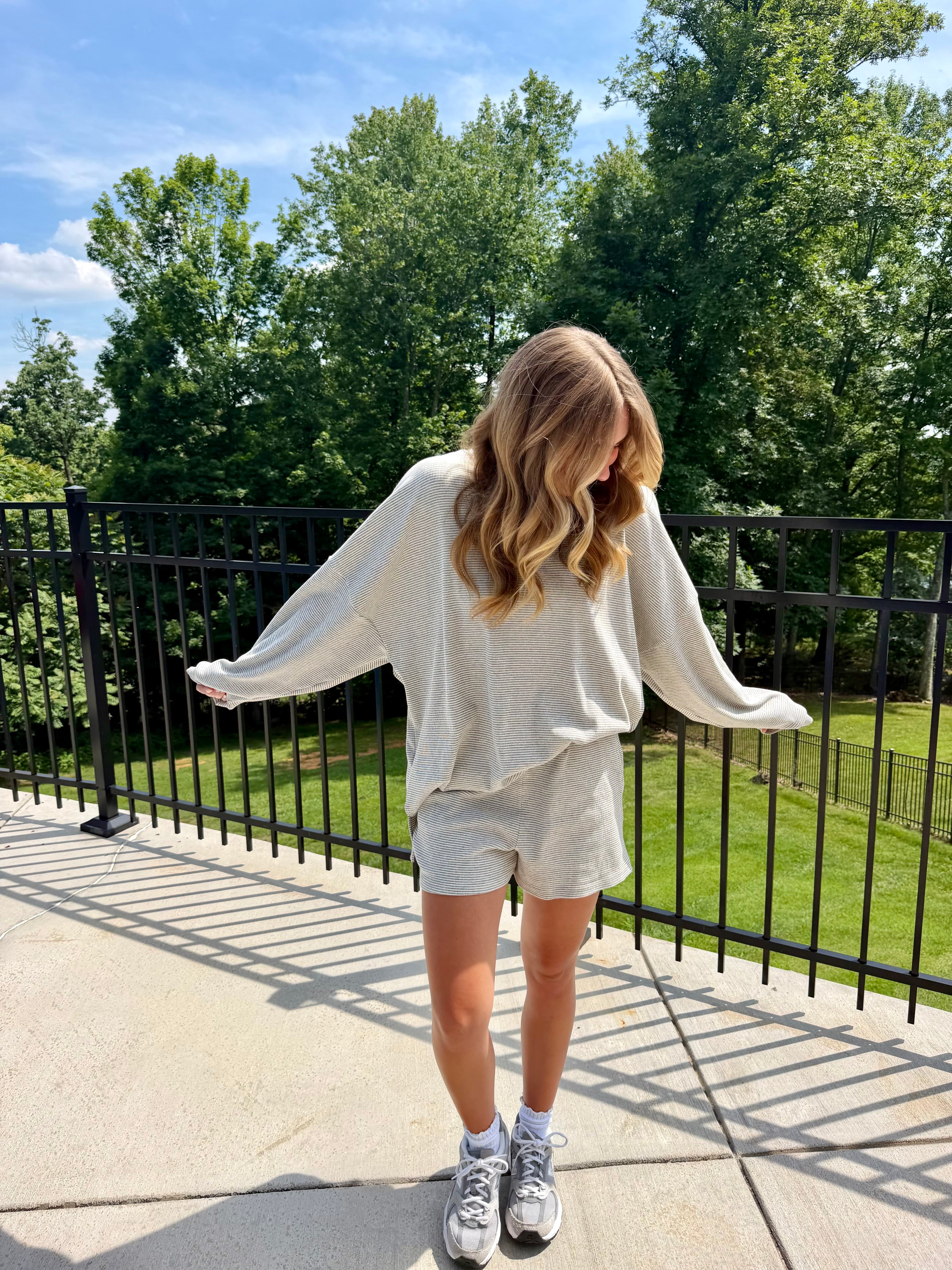 Person wearing a white long-sleeve shirt and gray shorts standing on a patio with trees in the background.