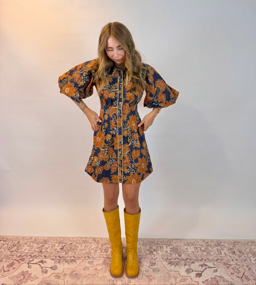 A woman stands against a plain light gray studio backdrop wearing a short floral dress with voluminous puff sleeves and a front zipper, paired with tall mustard yellow boots.