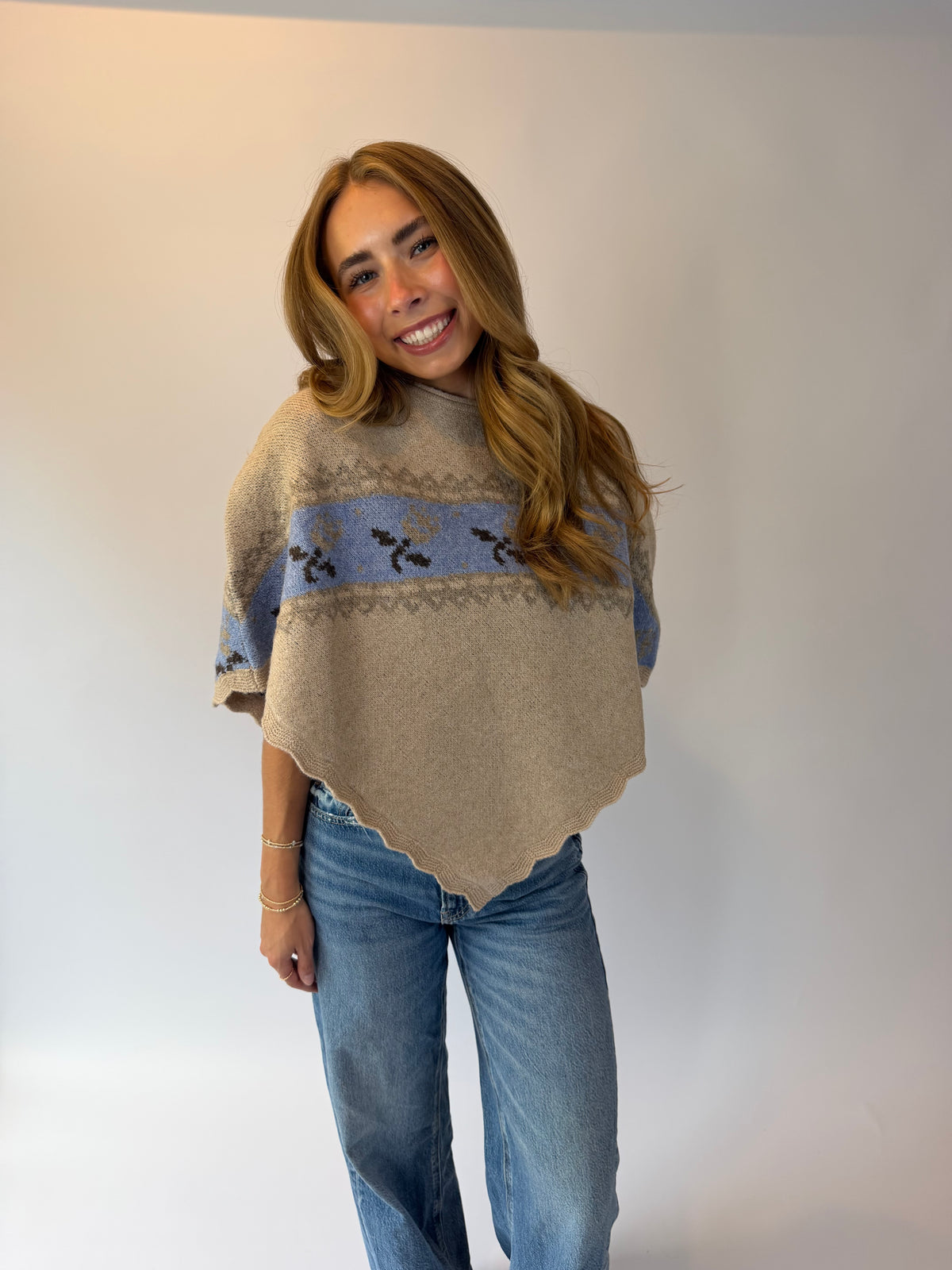 A woman modeling a beige knit poncho sweater with a blue and black patterned horizontal band, paired with blue jeans, standing against a plain light gray studio background.