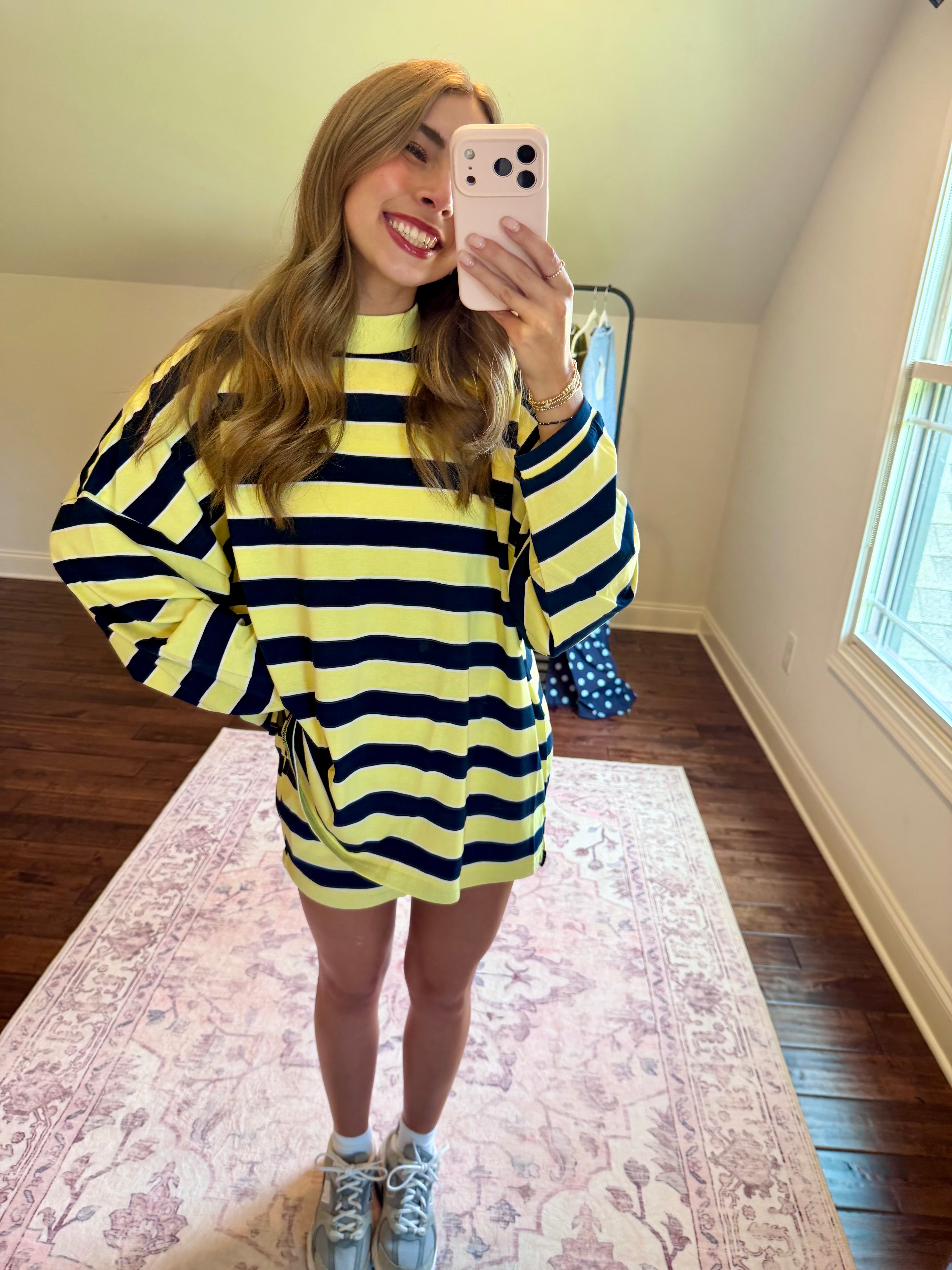 A woman takes a mirror selfie wearing a yellow and navy blue horizontally striped long-sleeve t-shirt dress. She is standing on a pink patterned area rug in a room with hardwood floors and a window.