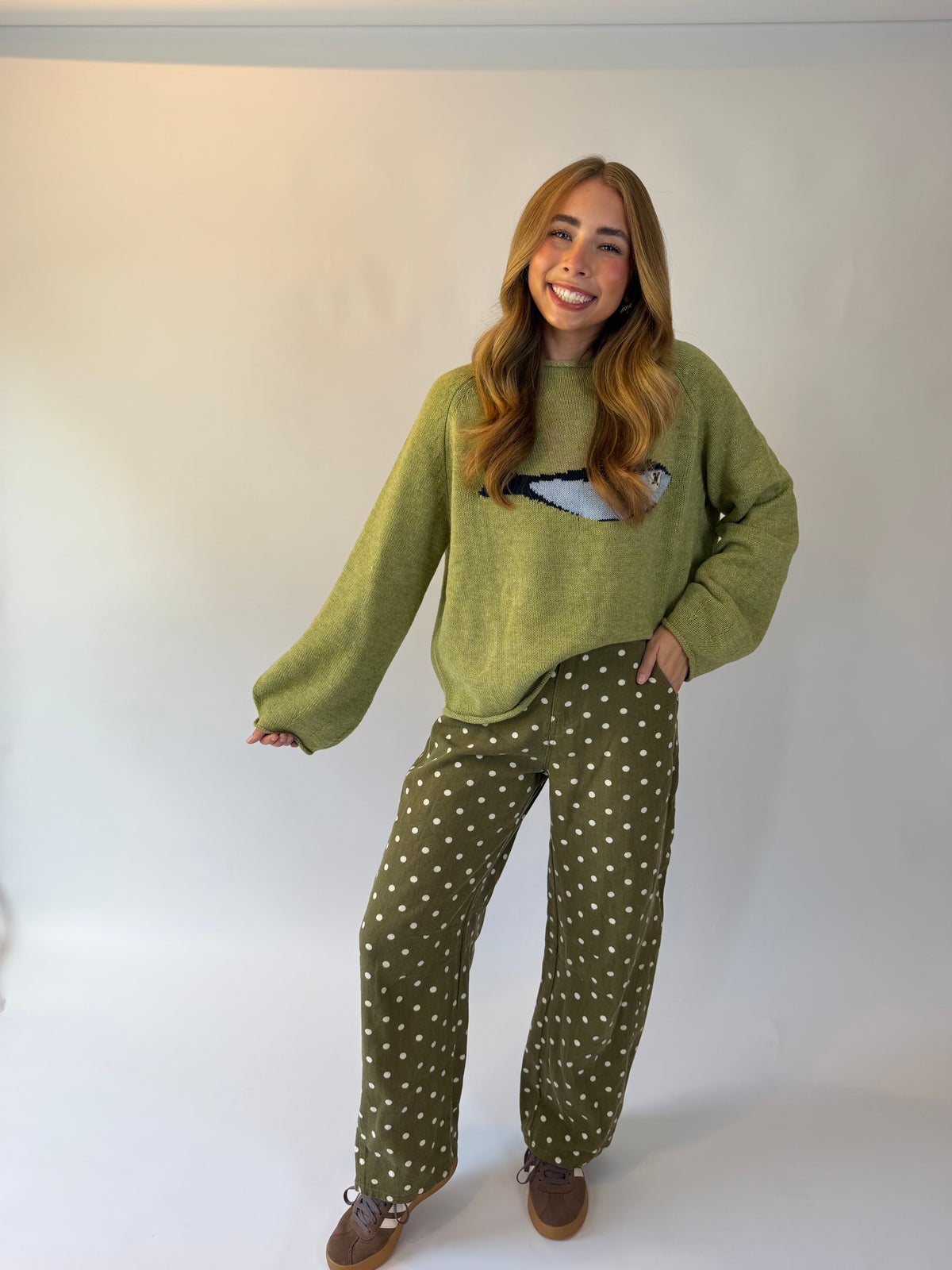A woman modeling a cozy two-piece knit loungewear set consisting of an olive green long-sleeve sweater with a white graphic on the chest and matching olive green pants with white polka dots, paired with brown shoes, against a plain white studio background.