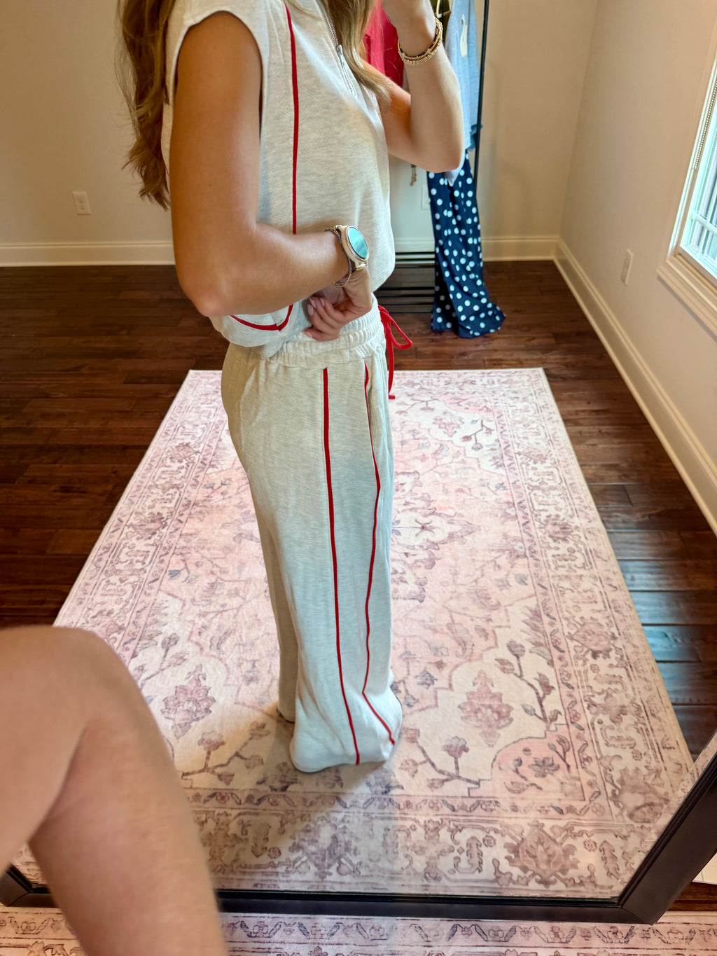 A woman stands in a room taking a mirror selfie to show off a matching two-piece lounge set. The set consists of a light beige sleeveless top and wide-leg pants, both featuring a vertical red stripe down the side.
