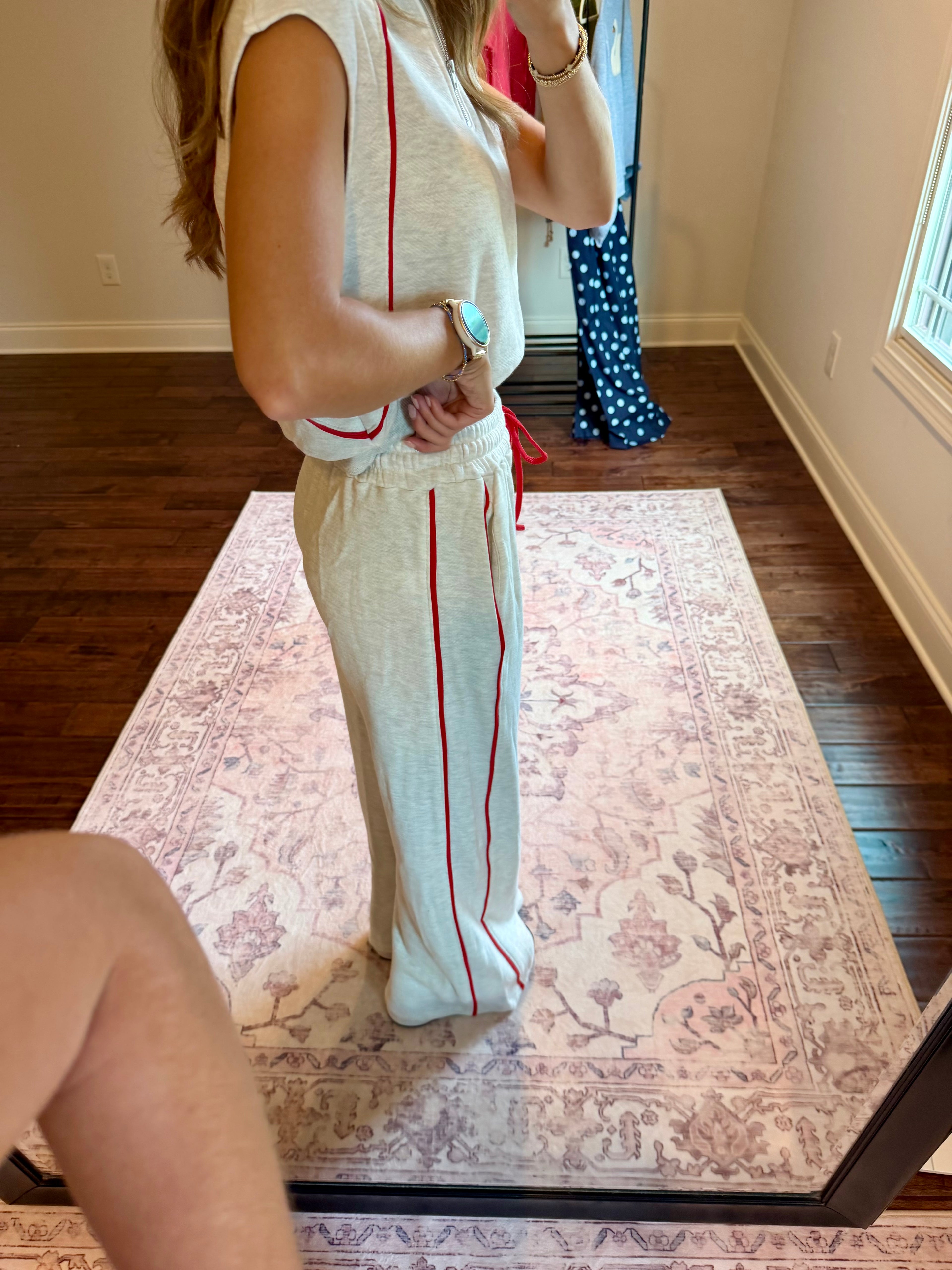 A woman stands in a room taking a mirror selfie to show off a matching two-piece lounge set. The set consists of a light beige sleeveless top and wide-leg pants, both featuring a vertical red stripe down the side.