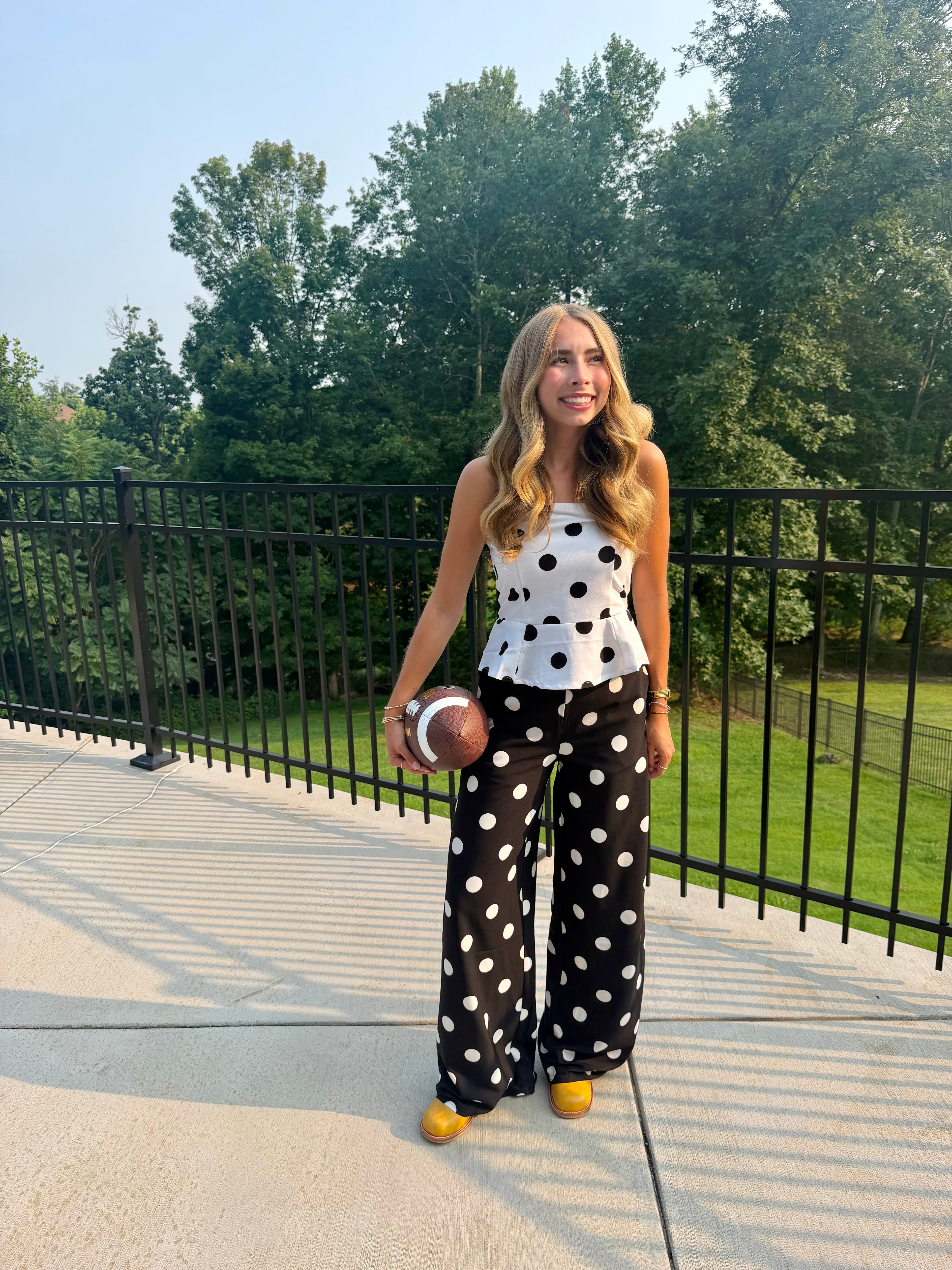 A woman stands outdoors on a concrete patio holding a football, wearing a white sleeveless top with black polka dots and matching wide-leg black polka dot pants, with yellow shoes. Trees and a black metal fence are behind her.