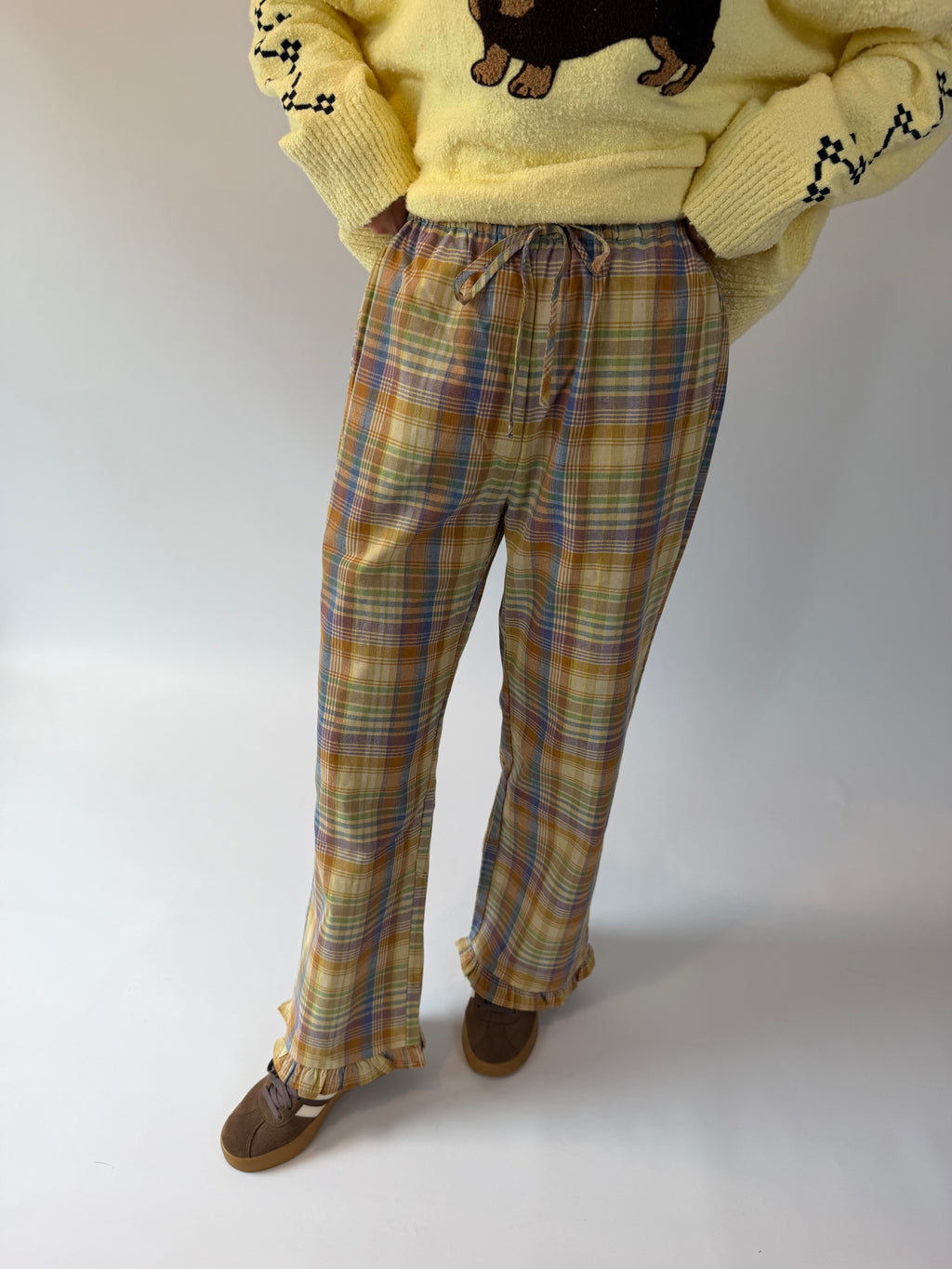 A person wearing plaid lounge pants in yellow, blue, and brown tones, paired with a pale yellow sweater and brown shoes, standing against a plain light background.