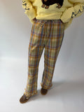 A person wearing plaid lounge pants in yellow, blue, and brown tones, paired with a pale yellow sweater and brown shoes, standing against a plain light background.