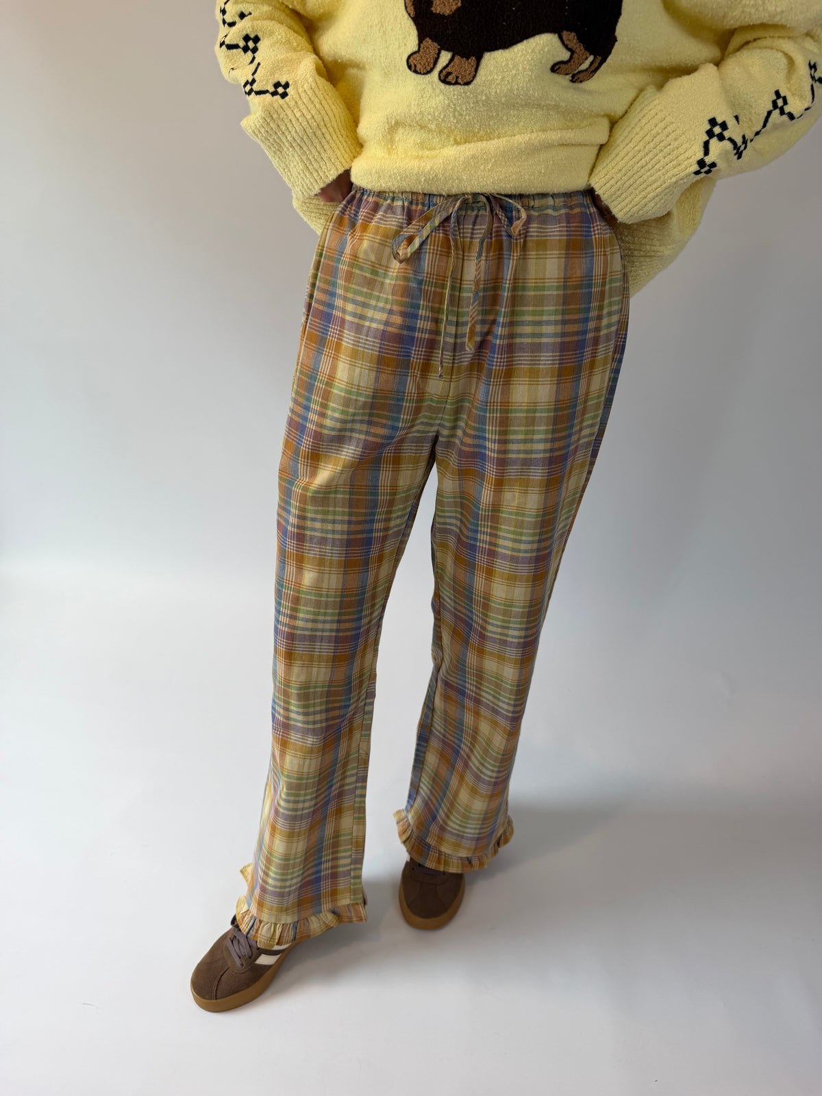 A person wearing plaid lounge pants in yellow, blue, and brown tones, paired with a pale yellow sweater and brown shoes, standing against a plain light background.
