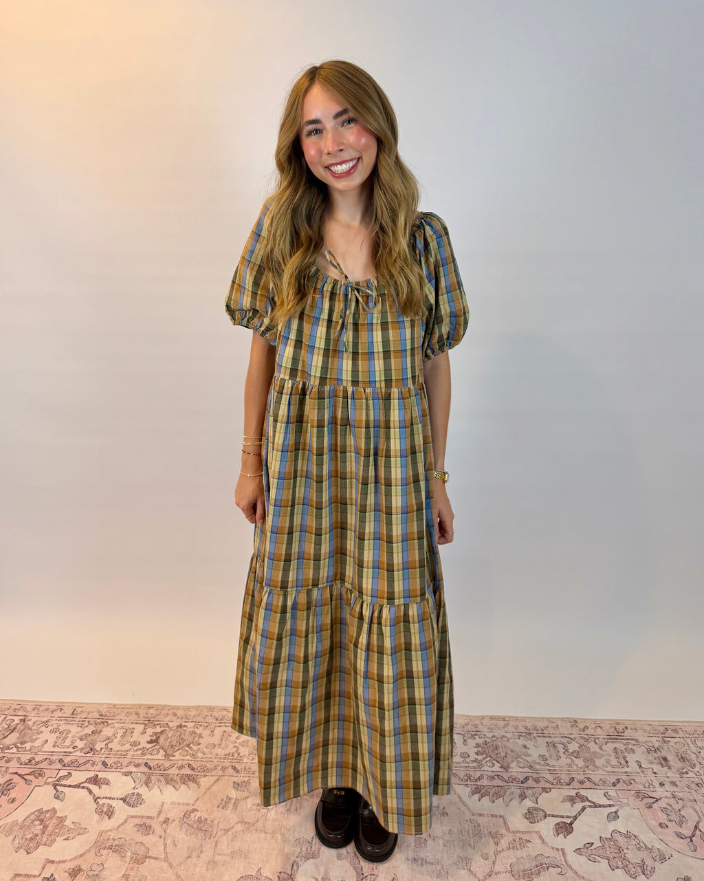 A woman stands against a plain light wall wearing a long-sleeved, tiered plaid maxi dress in yellow, green, and blue tones. She is smiling and posing, with a patterned rug visible underneath her.