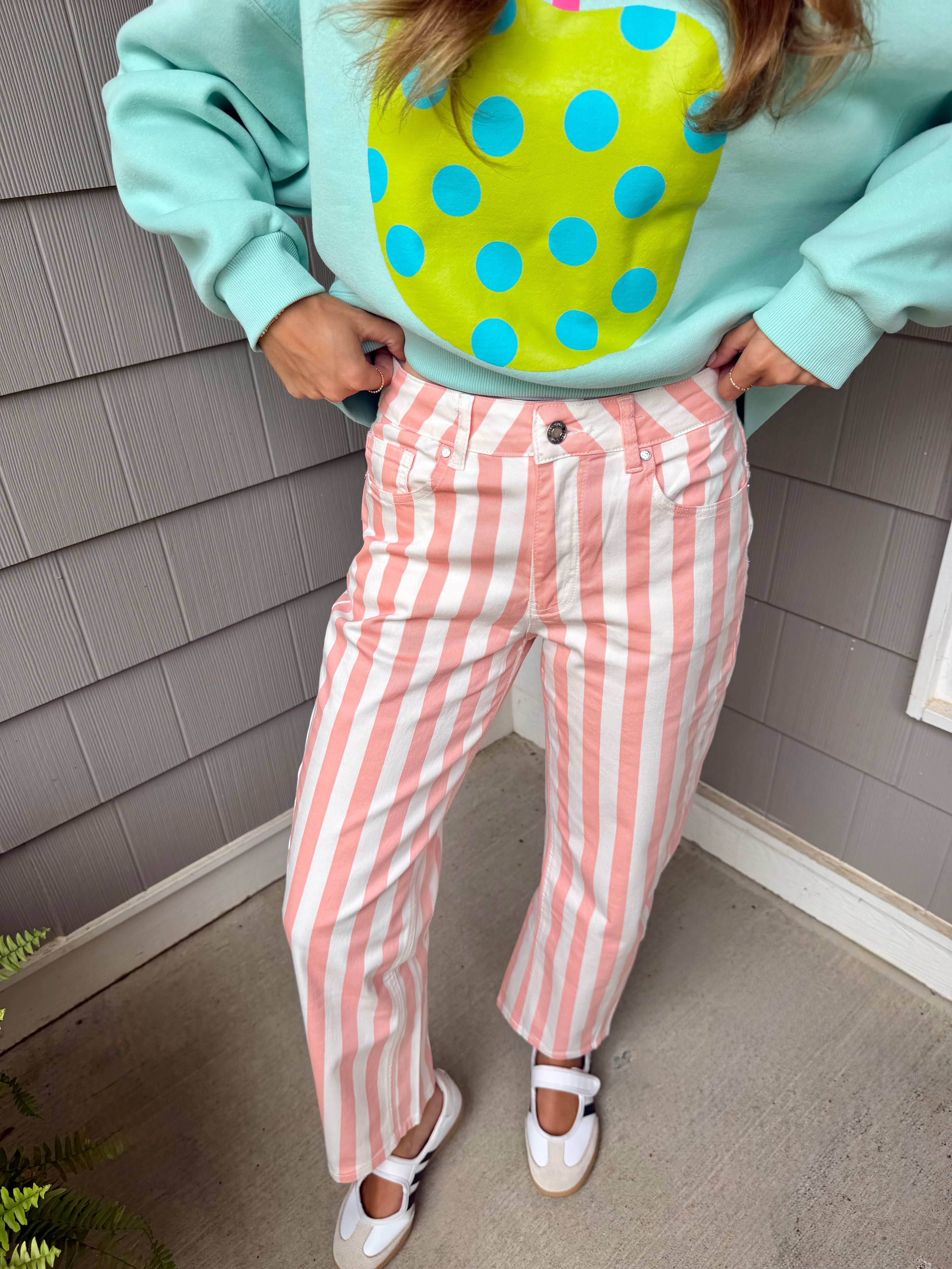 A person wearing pastel pink and white vertically striped jeans, a mint green sweatshirt with a yellow and blue polka dot graphic, and white Mary Jane shoes, standing against gray shingled house siding.